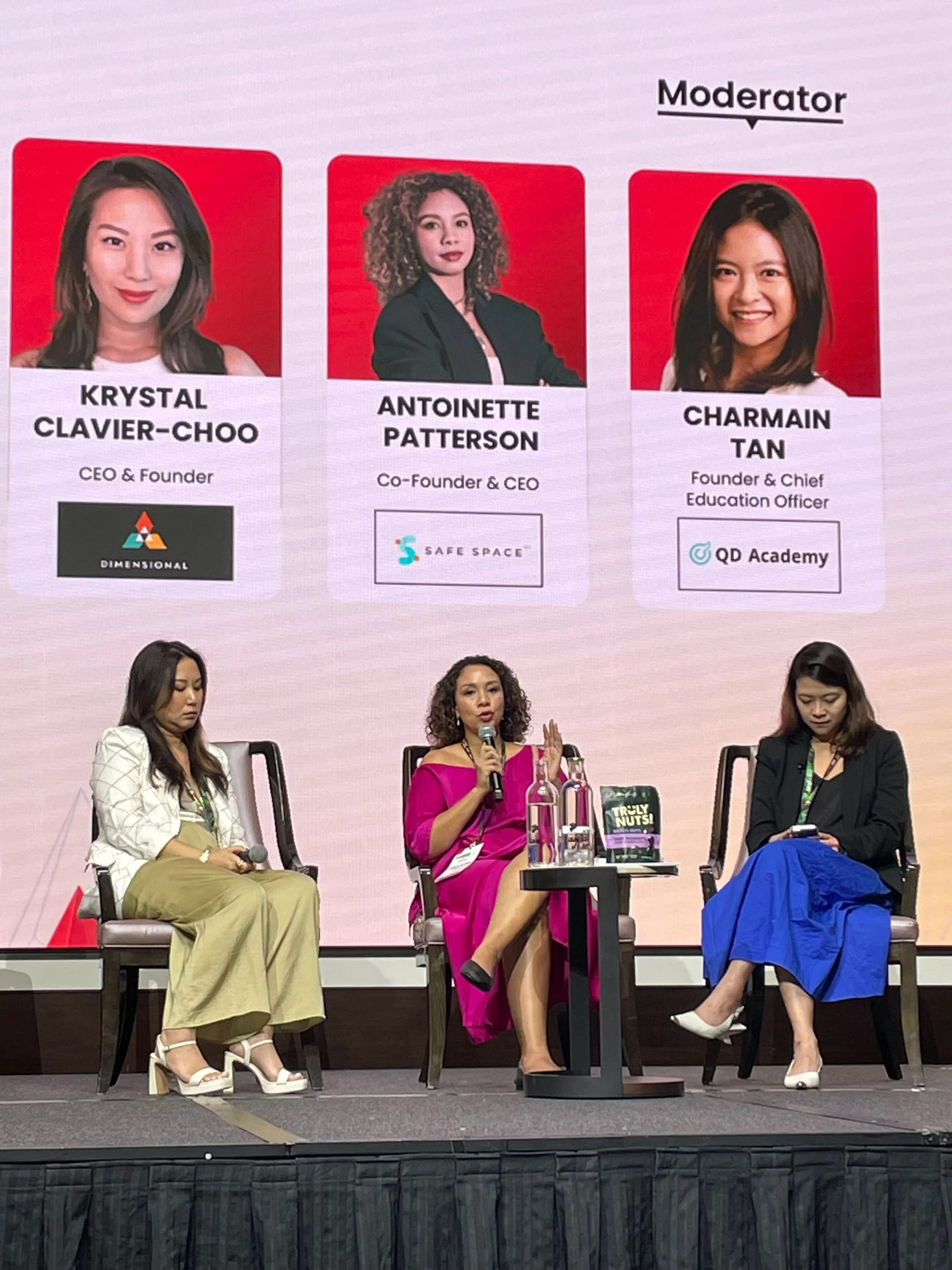 Panel discussion with three women seated on stage, with a large screen behind them showing headshots and titles of the speakers. The woman in the middle is speaking into a microphone, wearing a pink dress, while the women on her sides are listening. 