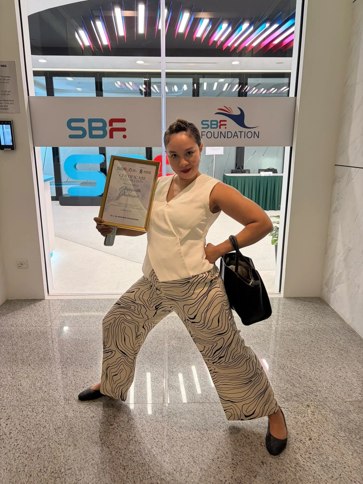 Woman standing in a confident pose with legs wide apart, holding a certificate in front of a glass door with the SBC Foundation logo, wearing beige patterned pants, a beige sleeveless top, and black shoes.