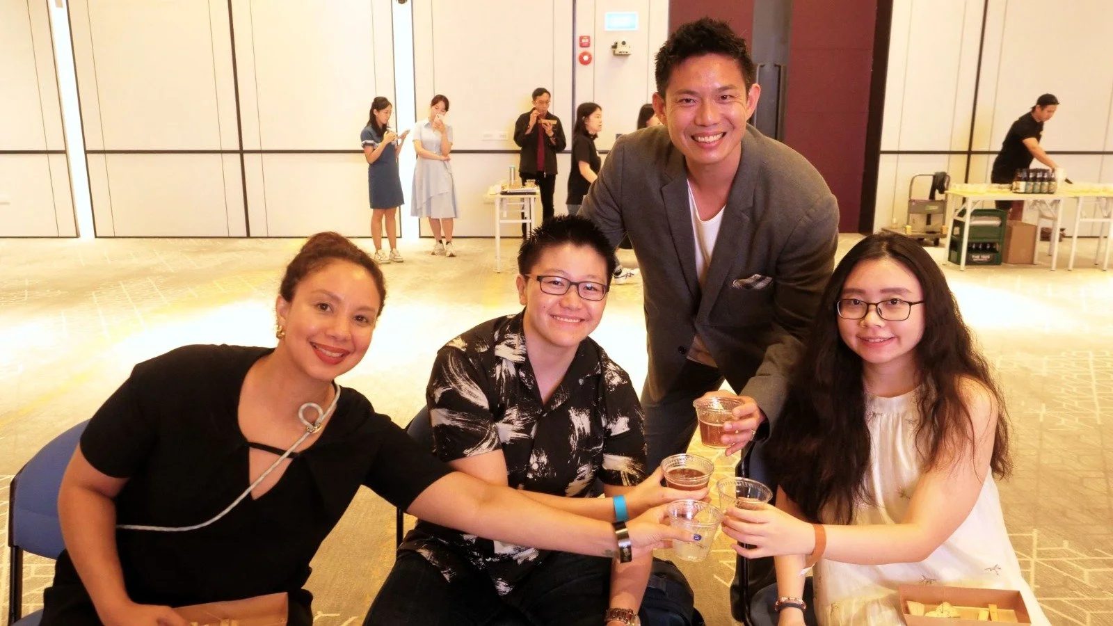 A group of four people sitting around a table at a social event, raising glasses of drinks for a toast, with a man standing behind them smiling. In the background, other people are standing and talking in a large, well-lit room.