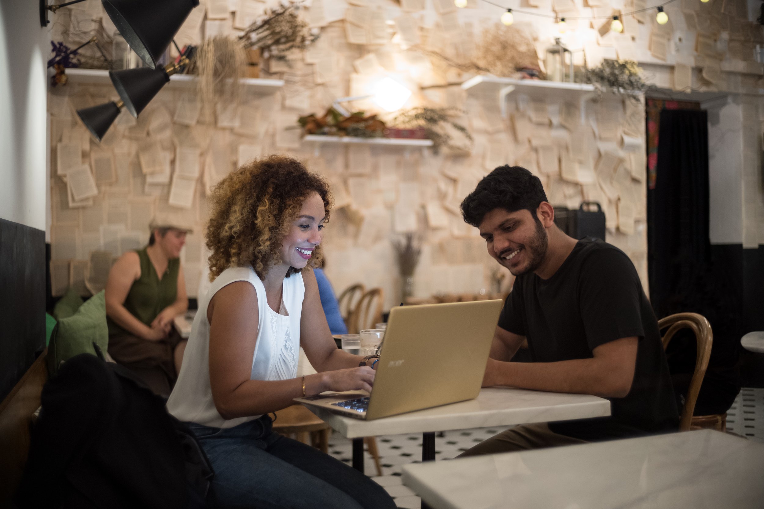 Two young adults, a woman with curly hair and a man with short dark hair, are sitting at a table in a cafe or restaurant, smiling and looking at a laptop in front of them. The background shows an artful wall decoration with books and branches, with other patrons in the background.