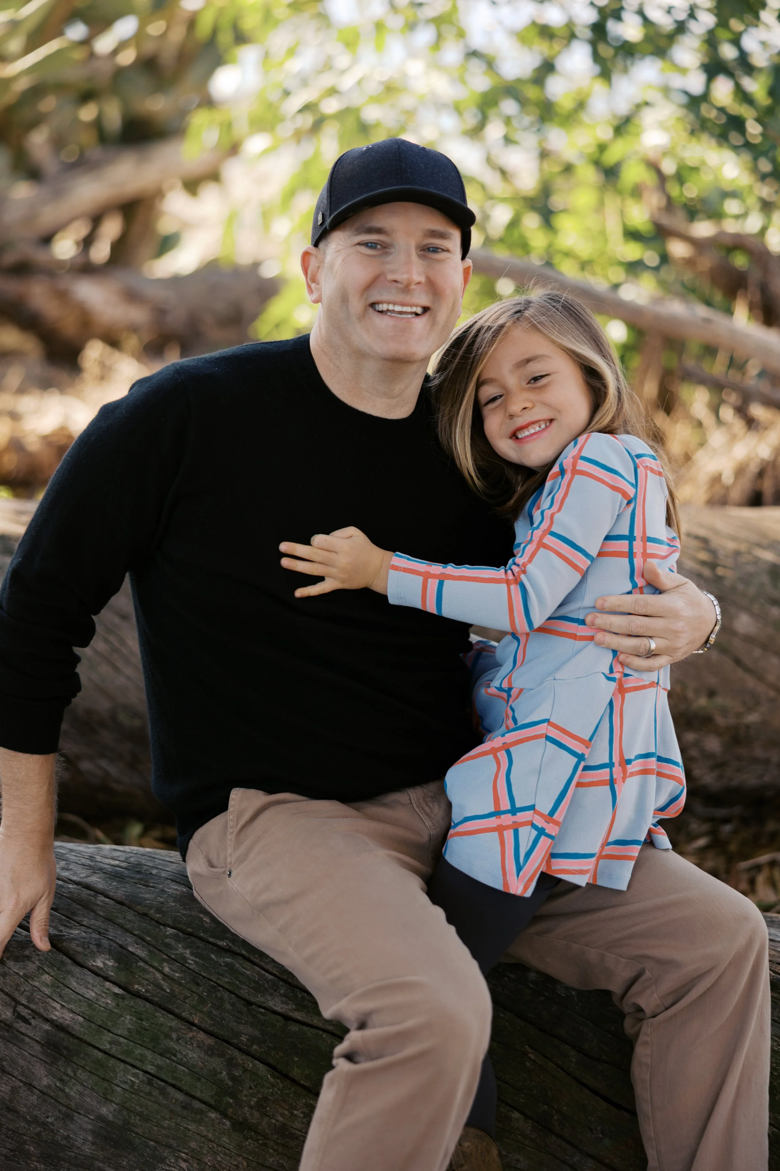 A man and a young girl sitting on a fallen tree in a forest, smiling and hugging each other.