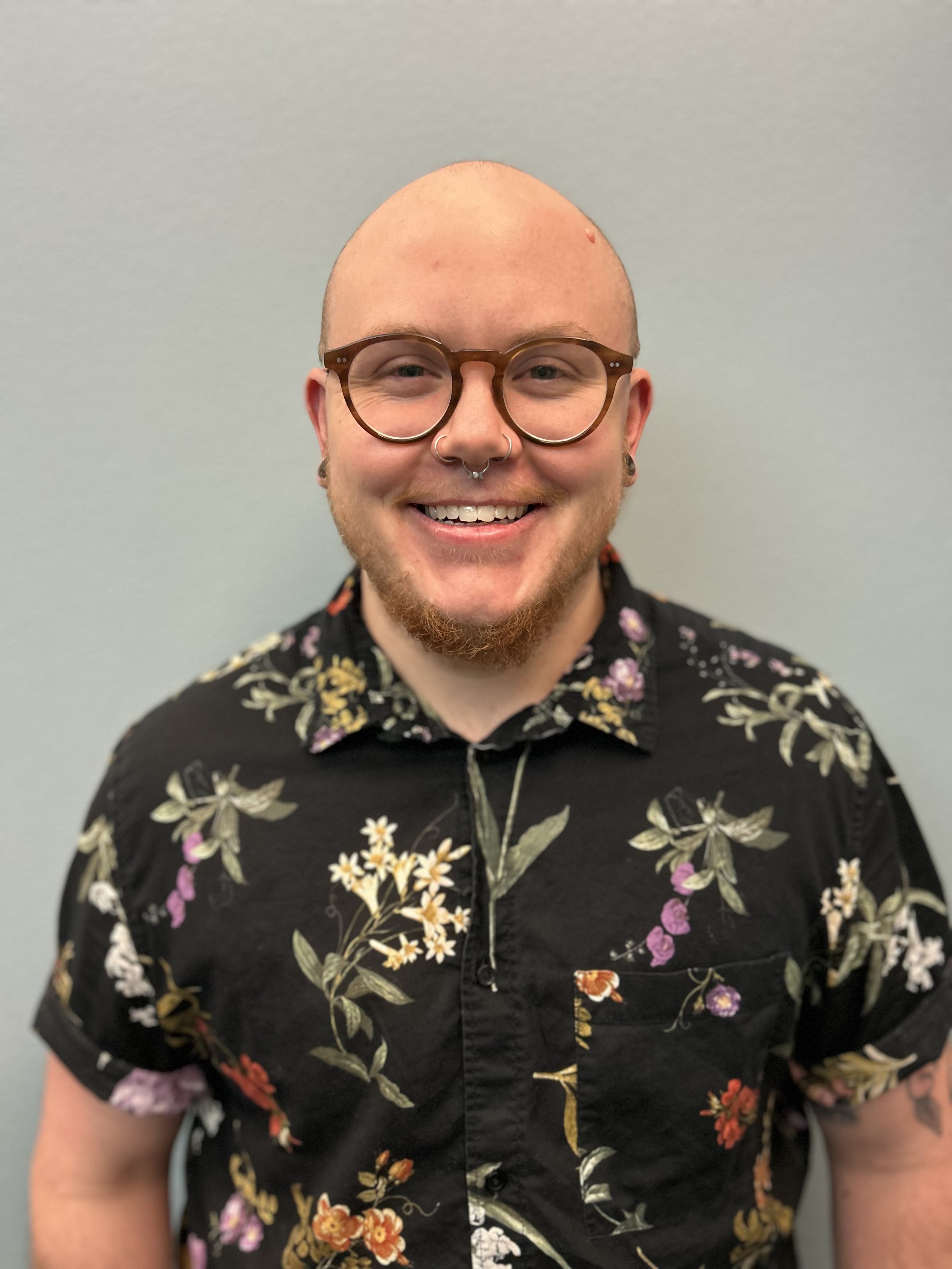 A smiling man with a bald head, glasses, and a septum piercing, wearing a black floral shirt.