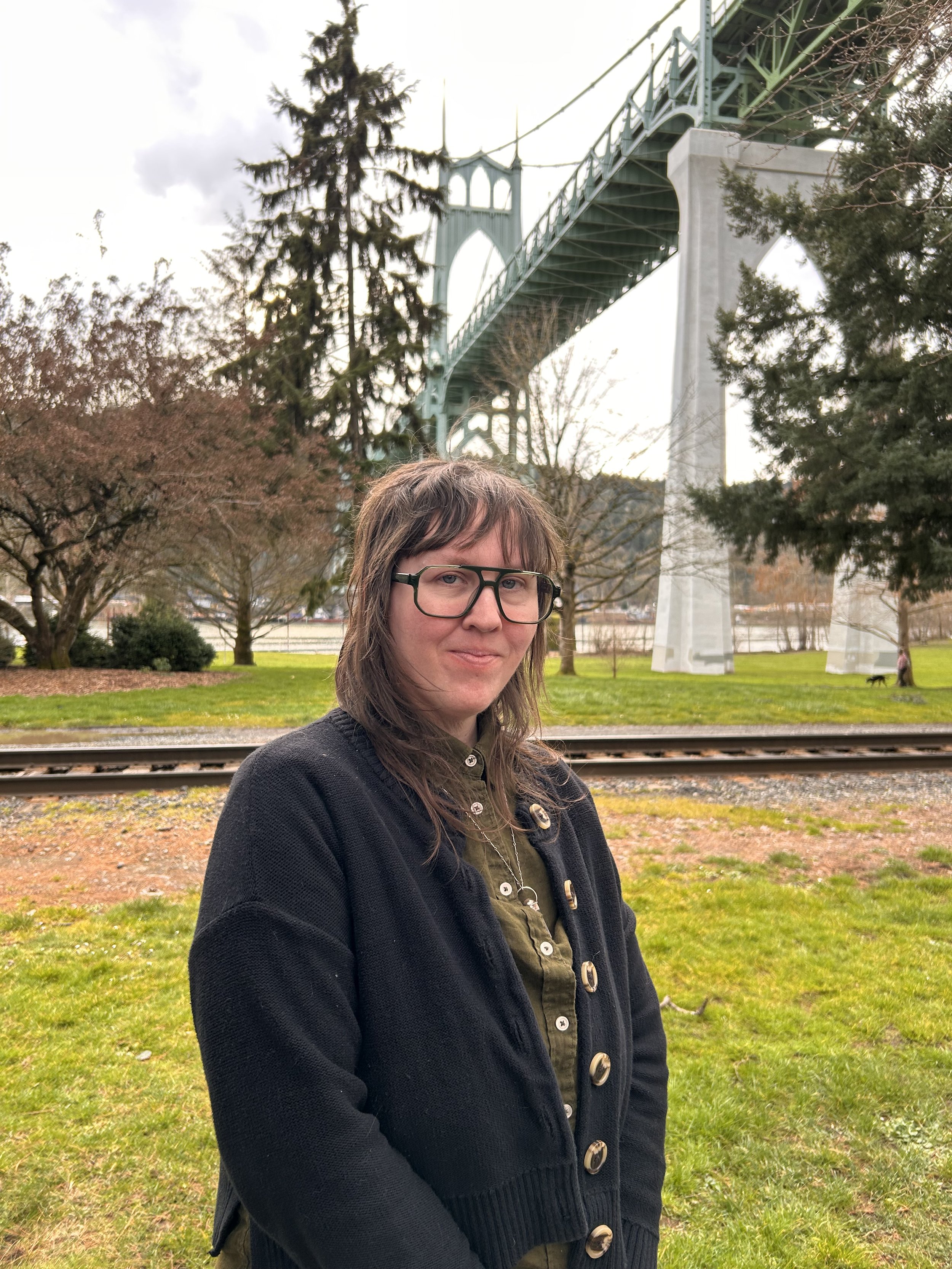 A woman with glasses and brown hair stands outdoors near railway tracks, with trees and a bridge in the background, during an overcast day.