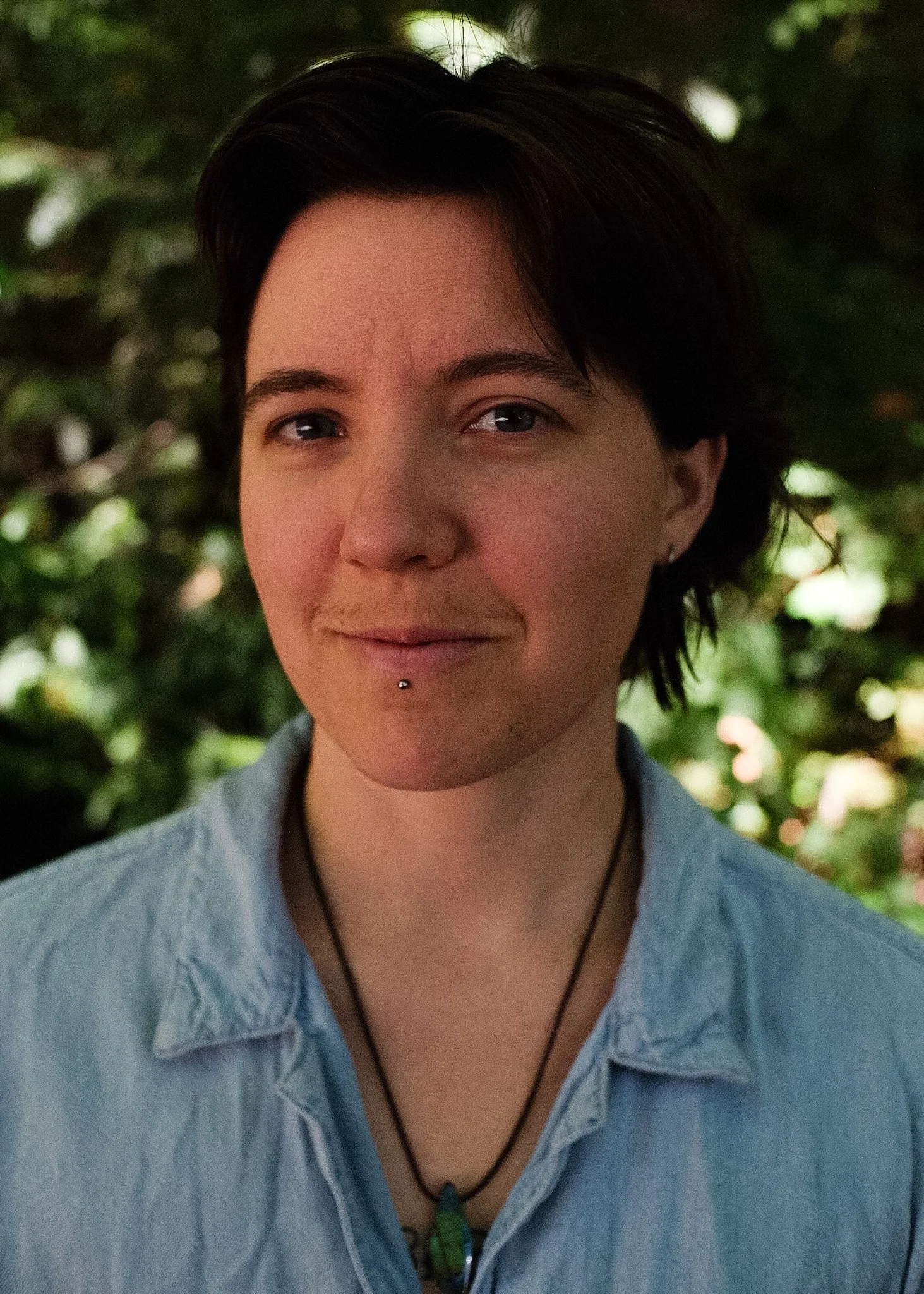 Close-up of a person with short dark hair, wearing a blue shirt, and a green pendant necklace, outdoors with blurred green foliage in the background.
