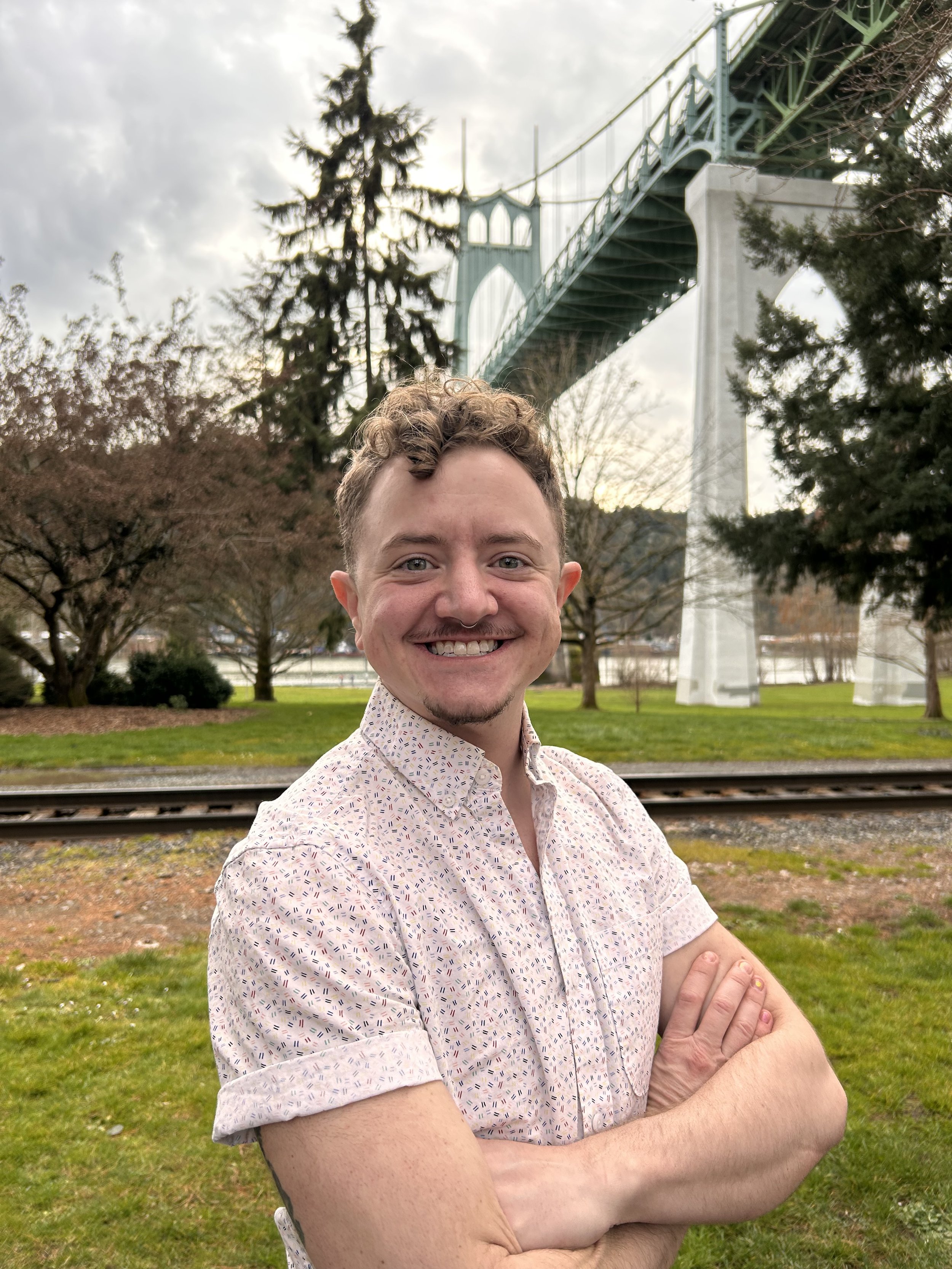 A young man with curly hair and a nose ring smiling at the camera with arms crossed, standing outdoors near train tracks, with trees and a bridge in the background.