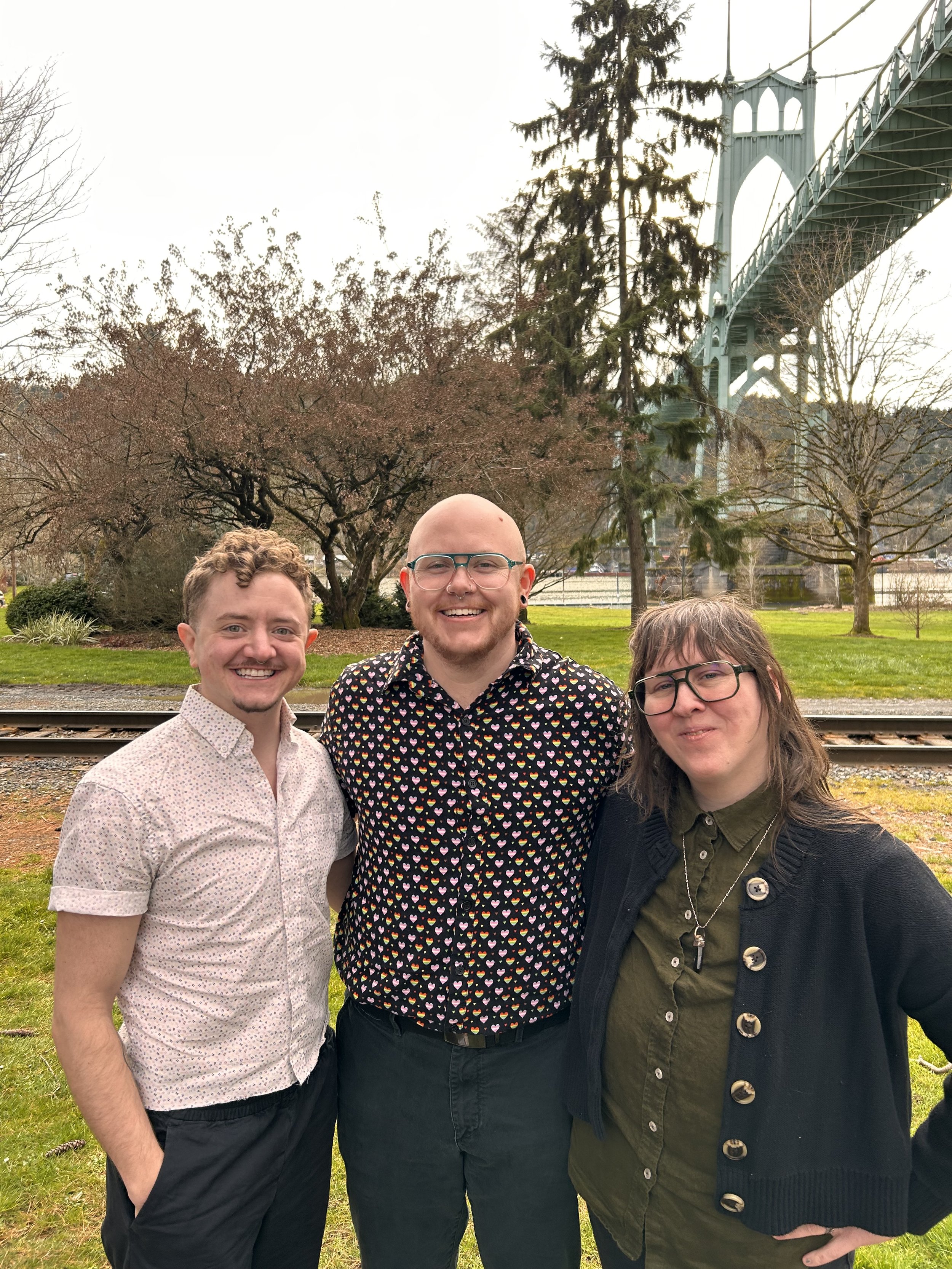 Three smiling friends standing outdoors near railroad tracks with trees and a bridge in the background.