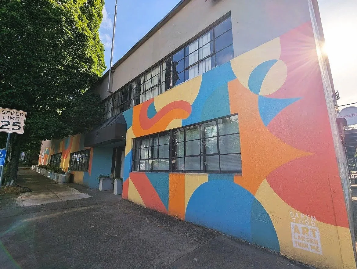 Colorful abstract mural on the side of a building with large windows, trees lining the sidewalk, and a speed limit sign of 25 mph. The mural includes various shapes in yellow, red, blue, and orange. Sunlight creates a lens flare effect in the top right corner.