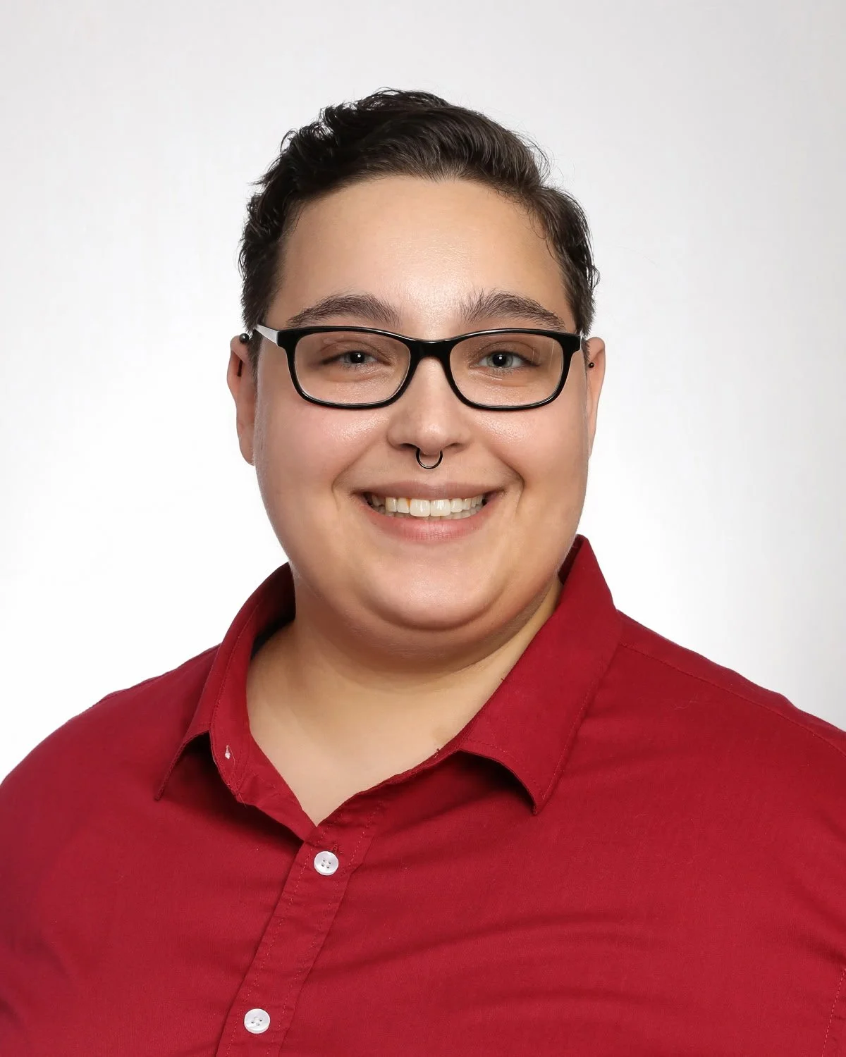 A person with short dark hair, glasses, and a septum piercing, smiling at the camera, wearing a red button-up shirt against a plain white background.