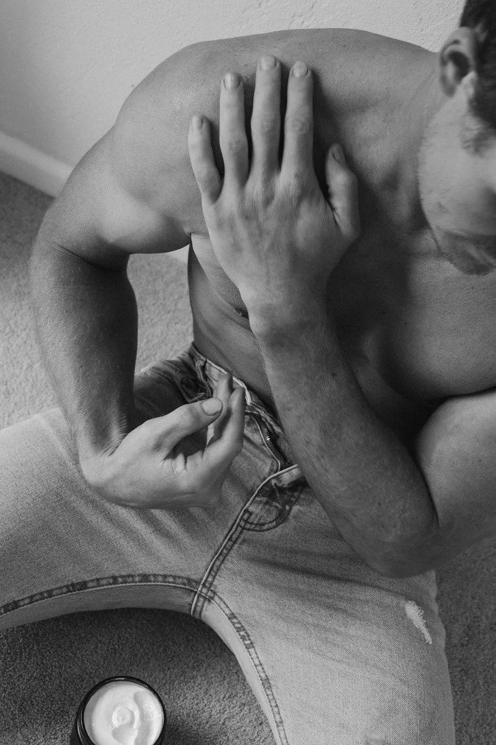 A monochrome photo of a shirtless man with a beard, sitting on a carpeted floor, pressing his hand against his shoulder, while another person's hand grasps his waist.