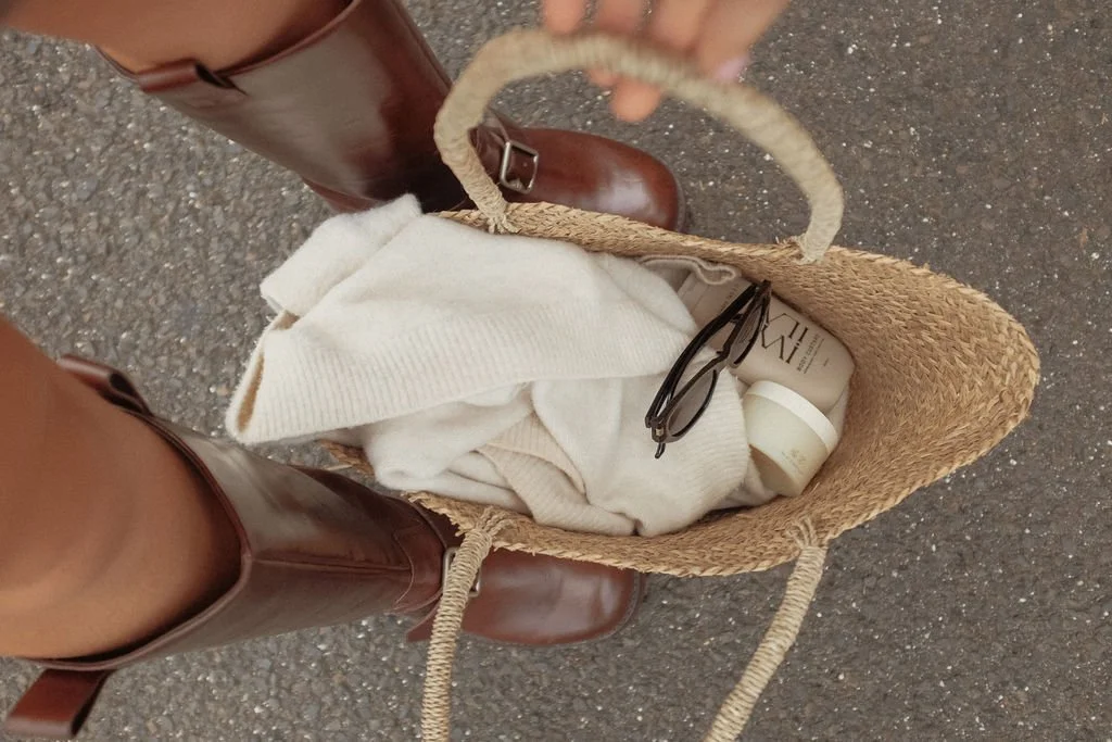A person standing on a paved surface wearing brown boots and a skirt, looking into a woven straw tote bag containing white clothing, a pair of sunglasses, and a cream-colored bottle.