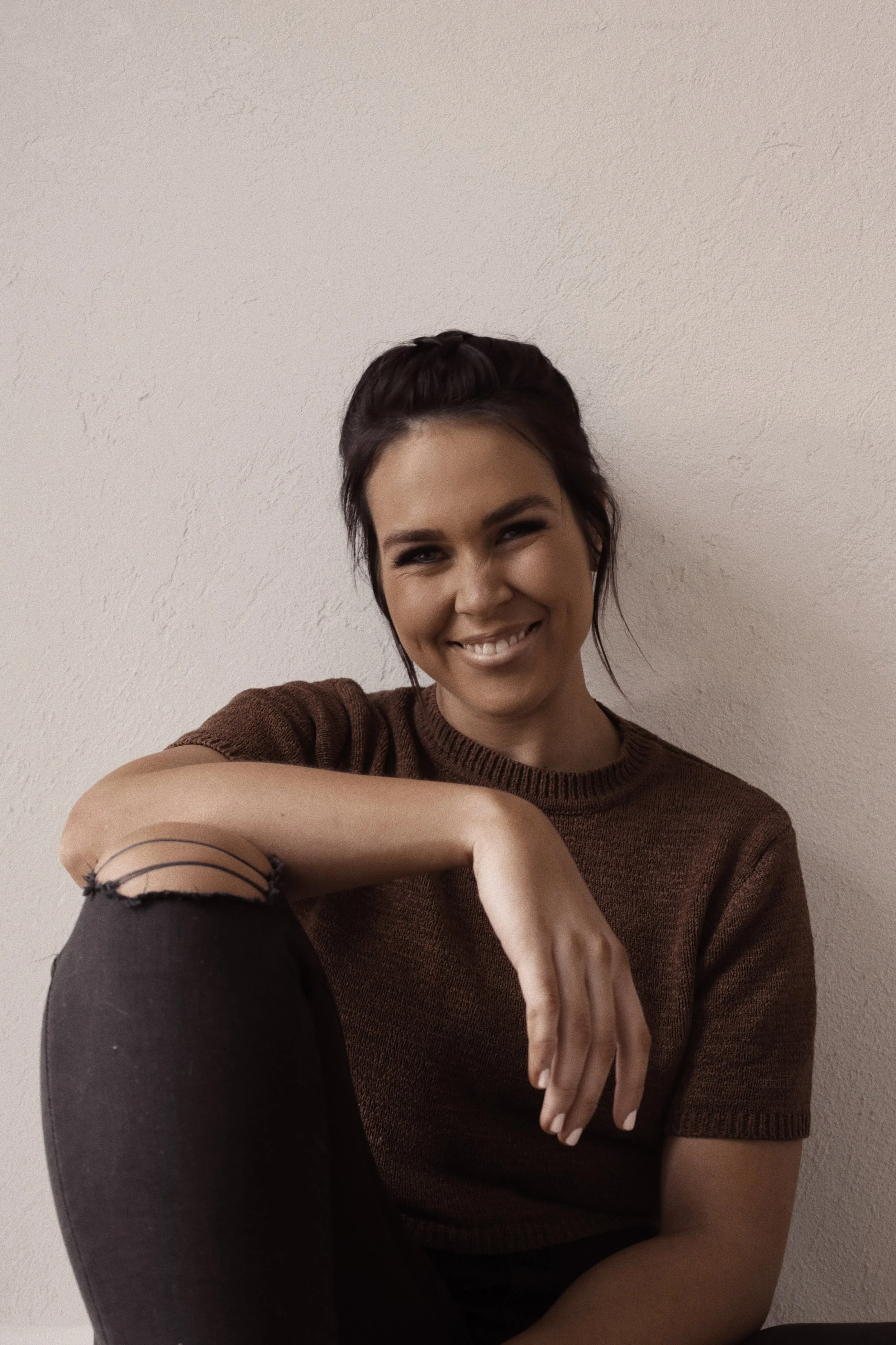 Smiling woman with dark hair pulled back, wearing a brown sweater and black ripped jeans, sitting against a plain beige wall.