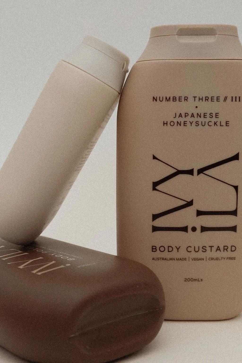 A beige bottle of Japanese honeysuckle body custard beside a white tube with a beige cap, and a brown soap dish or bar, all on a neutral background.