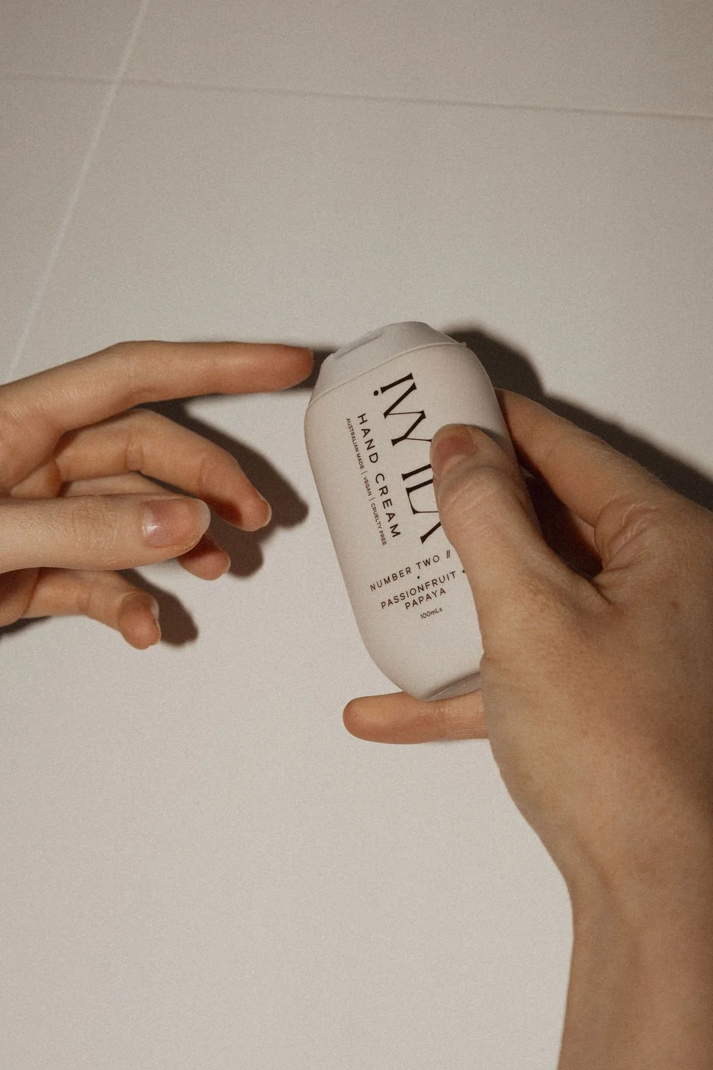 Person holding a white bottle of hand cream labeled 'IN MY' with a white tiled background.