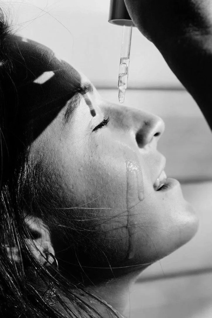 A close-up black and white photo of a woman with her eyes closed, receiving a liquid dropper treatment on her forehead from a person whose face is not visible.