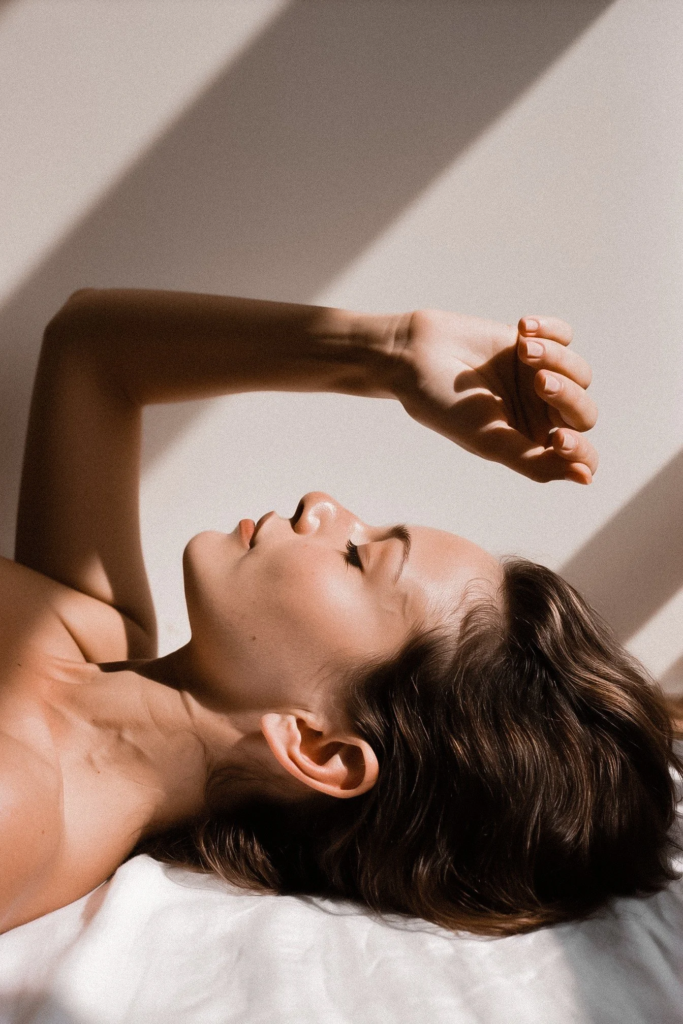 A woman lying on a white surface, with her eyes closed and a relaxed expression. Her hair is wavy and brown, and her hand is raised near her face. Shadows and soft lighting highlight her features.