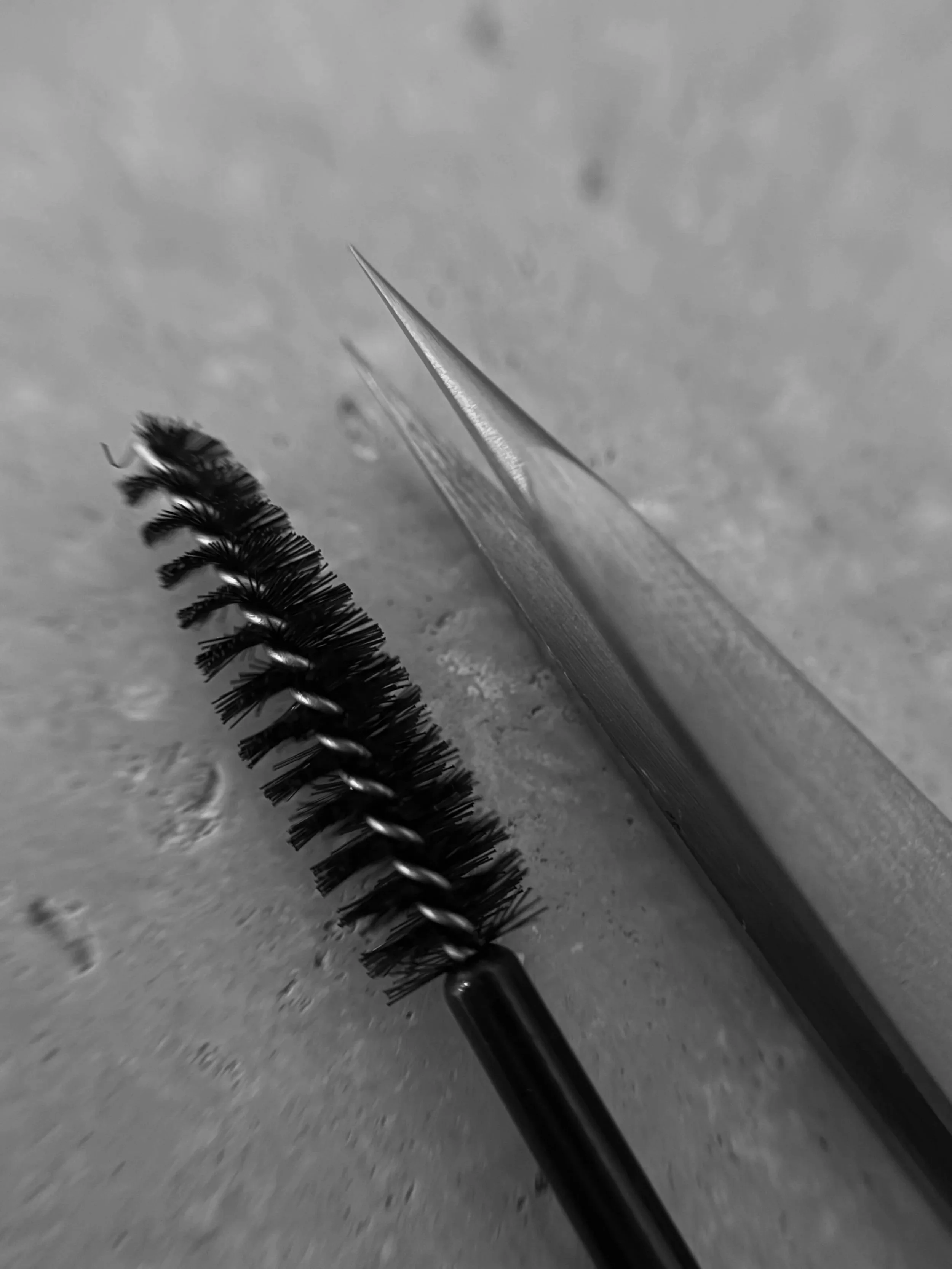 Close-up of a mascara wand with black mascara on bristles, and a spoolie brush, both lying on a gray textured surface.