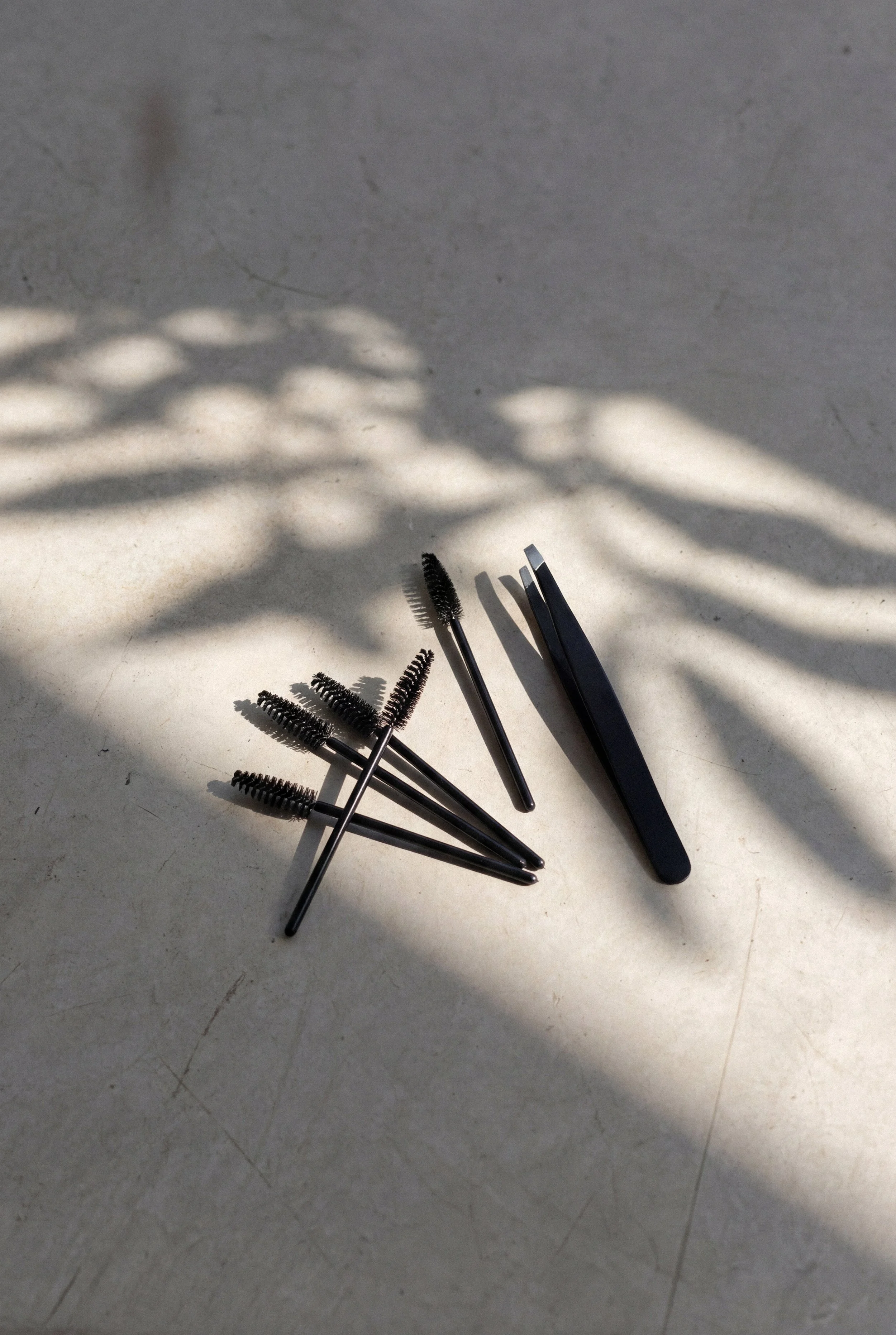 Several black eyelash brushes and a black eyelash curler on a light-colored surface with shadows of leaves.