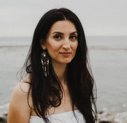 Woman with long dark hair standing on a beach with water and cloudy sky in the background.