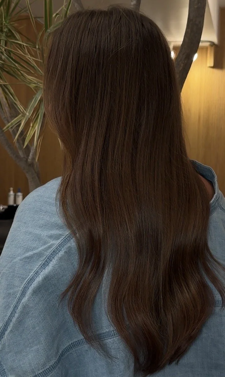 Back view of a woman with long, wavy, brown hair wearing a denim jacket indoors with a plant and wooden wall in the background.