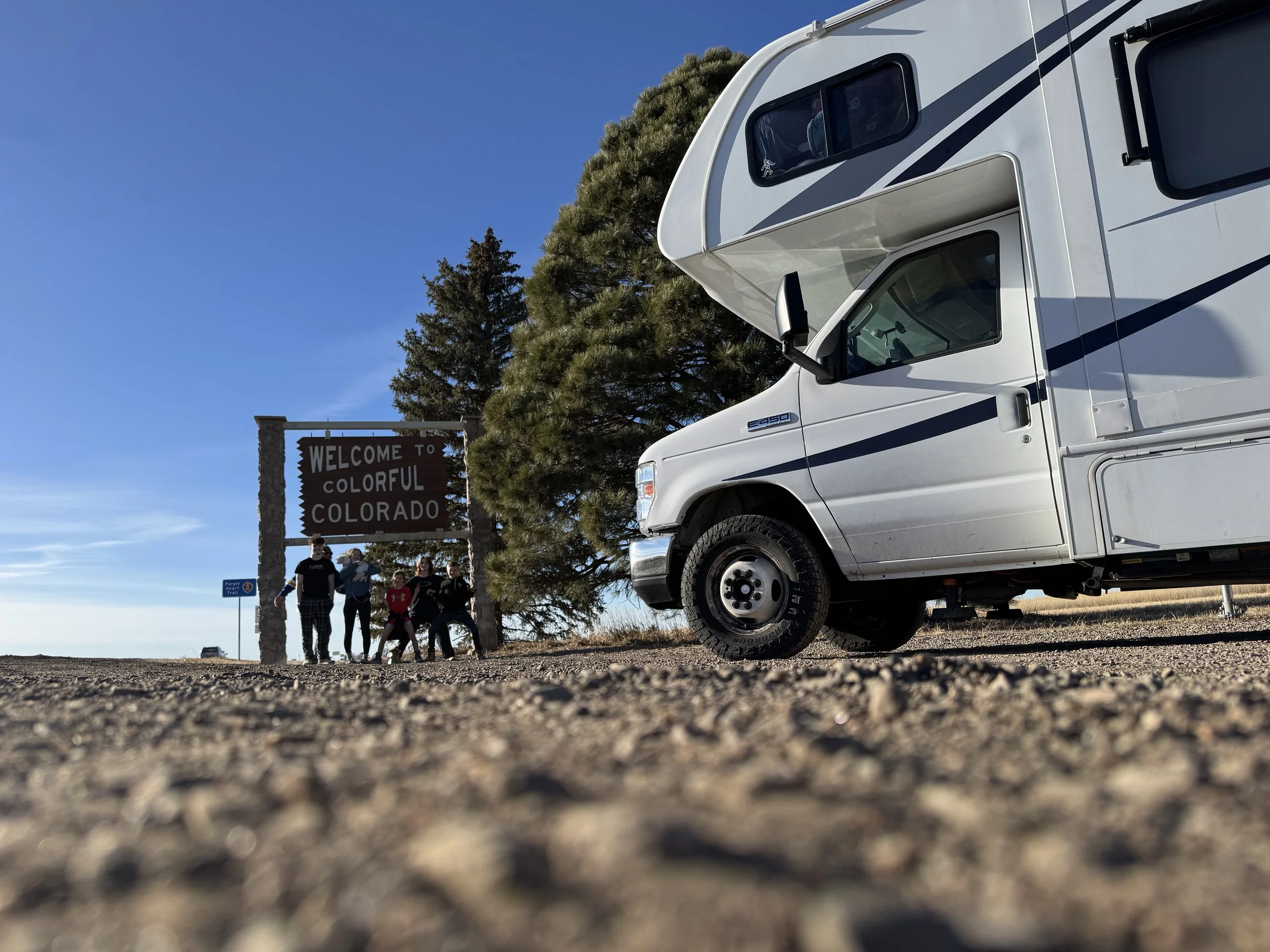 Group of people standing near a welcome sign that reads 'Welcome to Colorful Colorado', with a large white RV, trees, and a clear blue sky in the background.