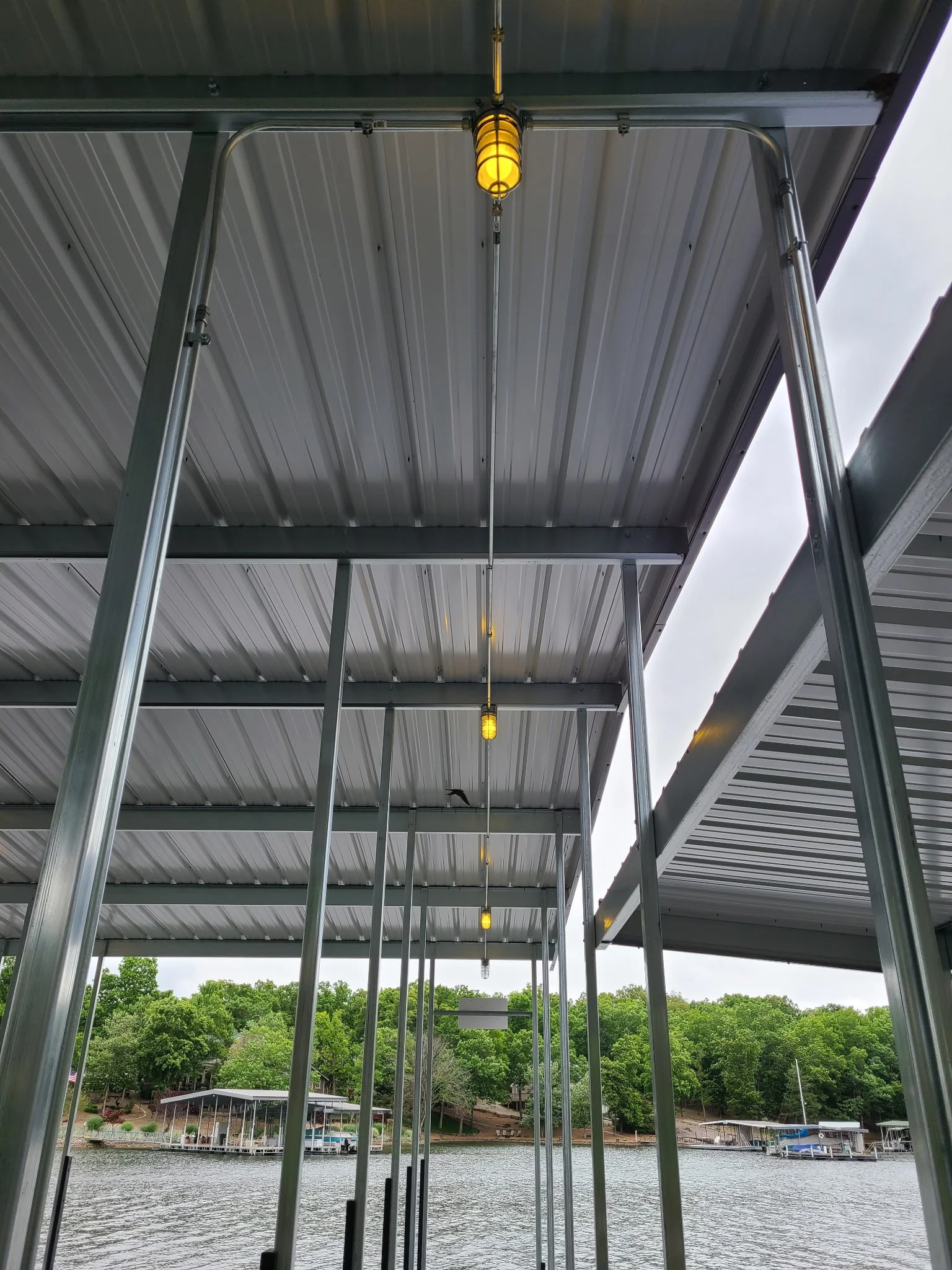 View of a marina with boats, water, trees, and a cloudy sky, seen from underneath a metallic structure with hanging yellow lights.