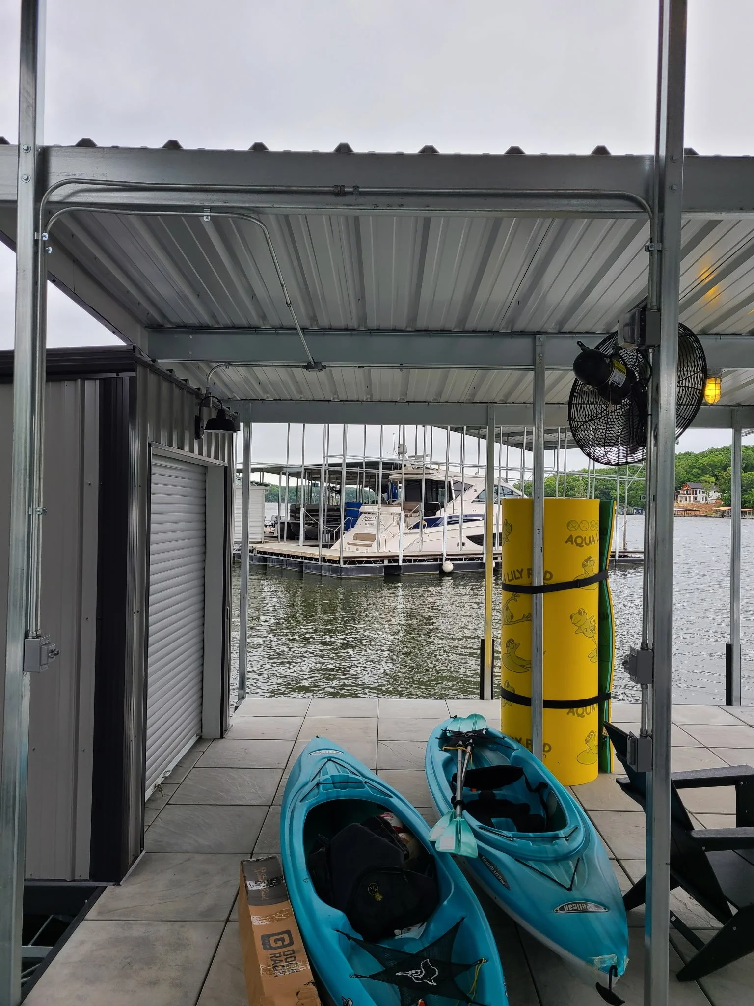 View of a marina dock with two blue kayaks in the foreground, a yellow floatation device labeled 'Aqua' on the right, and a boat docked in the water in the background, seen through a metal and glass structure.