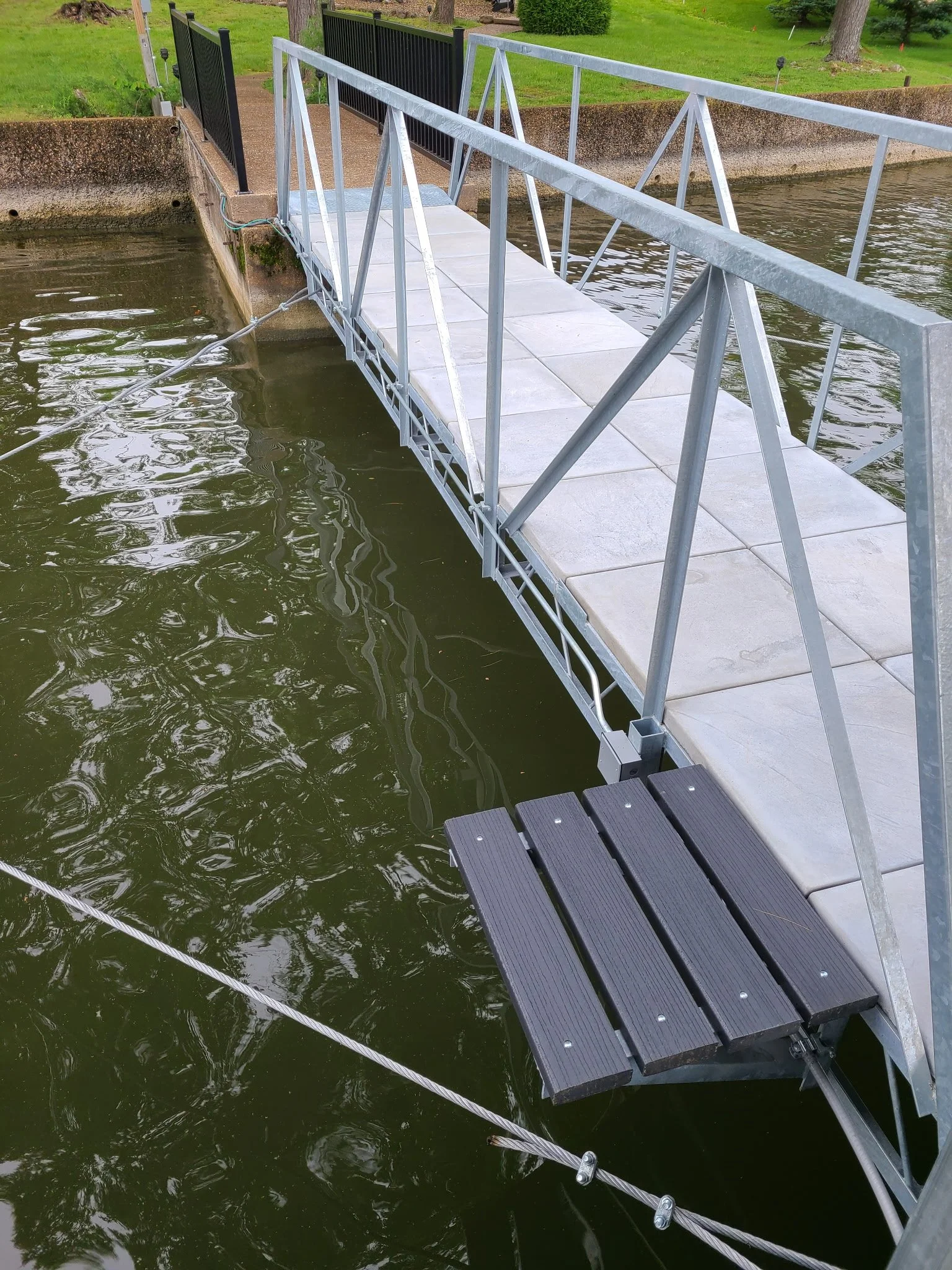 A metal and concrete boat ramp extending into a body of water, with a black railing at the top and a metal footbridge platform with a rubberized surface. There is grass and trees in the background.