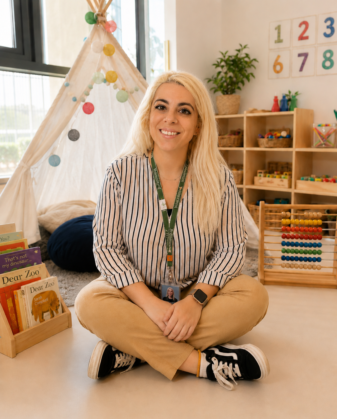 A young woman with blonde hair sitting cross-legged on the floor of a classroom or library, smiling at the camera. Behind her are shelves with children's books, toys, and educational materials, and a teepee decorated with colorful pom-poms. She is wearing a striped shirt, tan pants, and a smartwatch, and has an ID badge around her neck.
