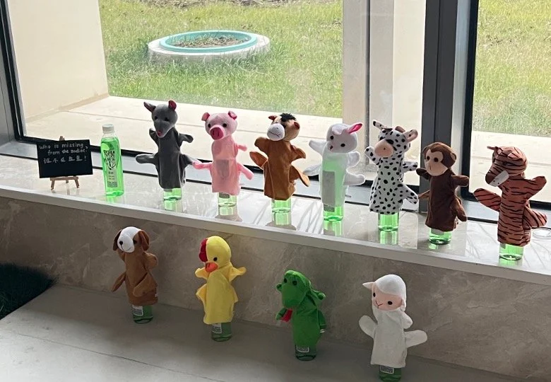 A collection of plush animal toys lined up on a windowsill and a lower surface, with a glass window showing a grassy outdoor area and a bird bath outside. The toys include various animals such as mice, pigs, a cow, a zebra, a monkey, a dog, a duck, a crocodile, and a sheep.