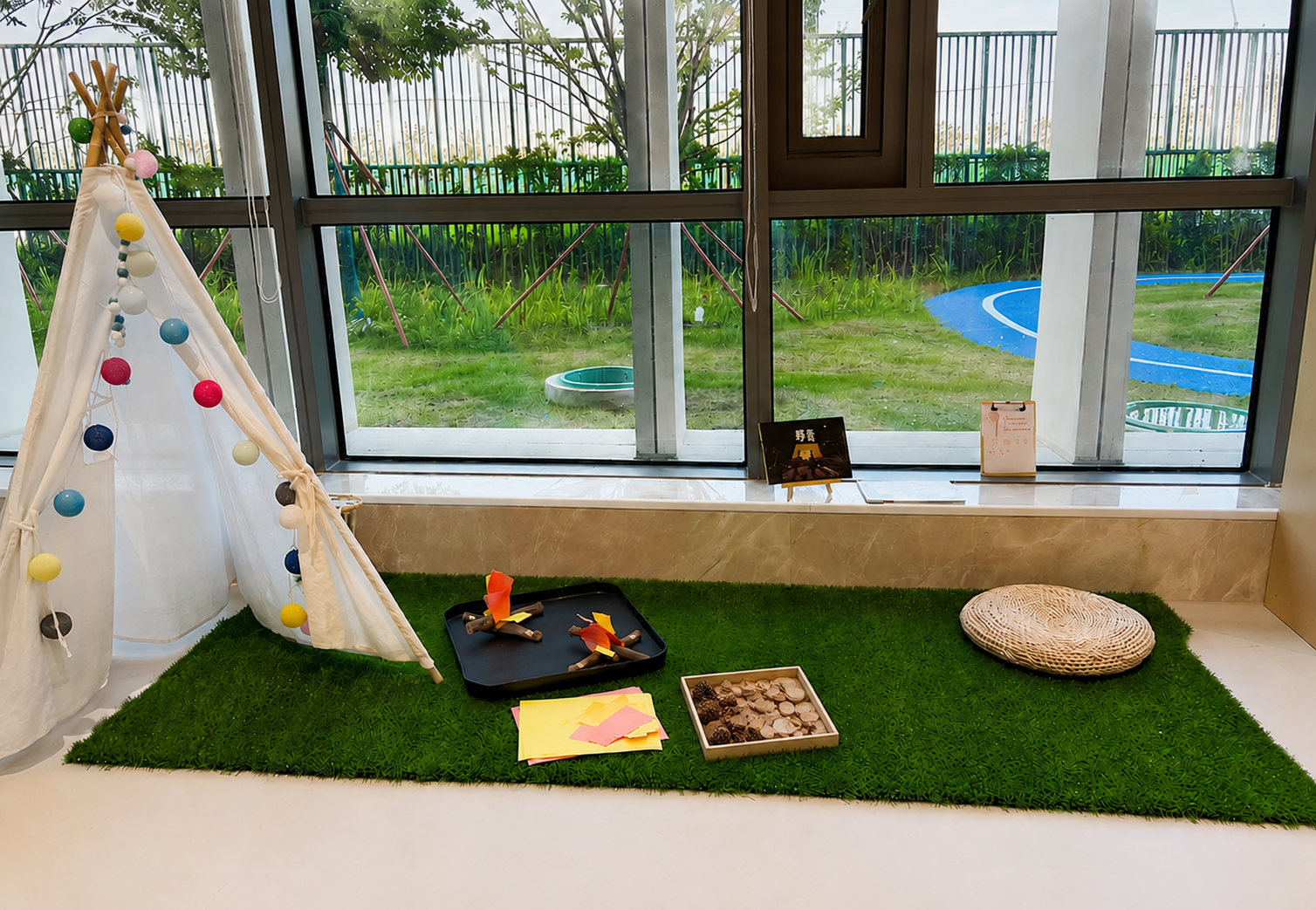 Indoor play area with a green artificial grass rug, a small teepee tent decorated with colorful balls, a tray with toy animals, some papers, and natural elements, a wicker cushion, and large glass windows overlooking a grassy outdoor space with a slide and garden.