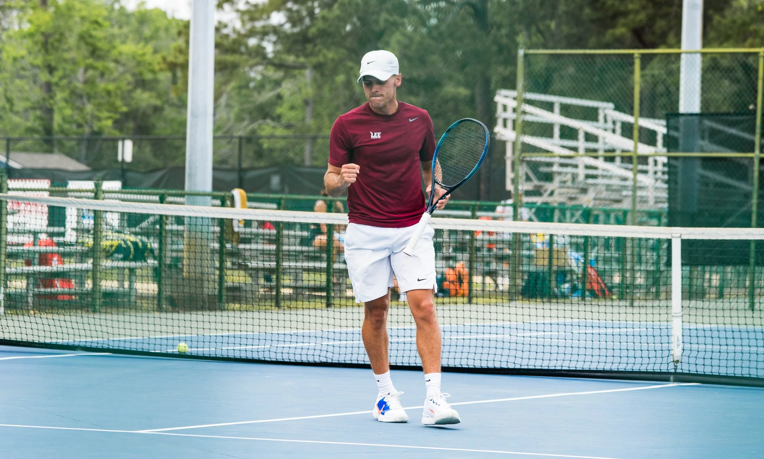 A tennis player in a maroon shirt, white shorts, and a white cap standing on a blue tennis court holding a tennis racket, with a tennis ball on the ground near his feet.