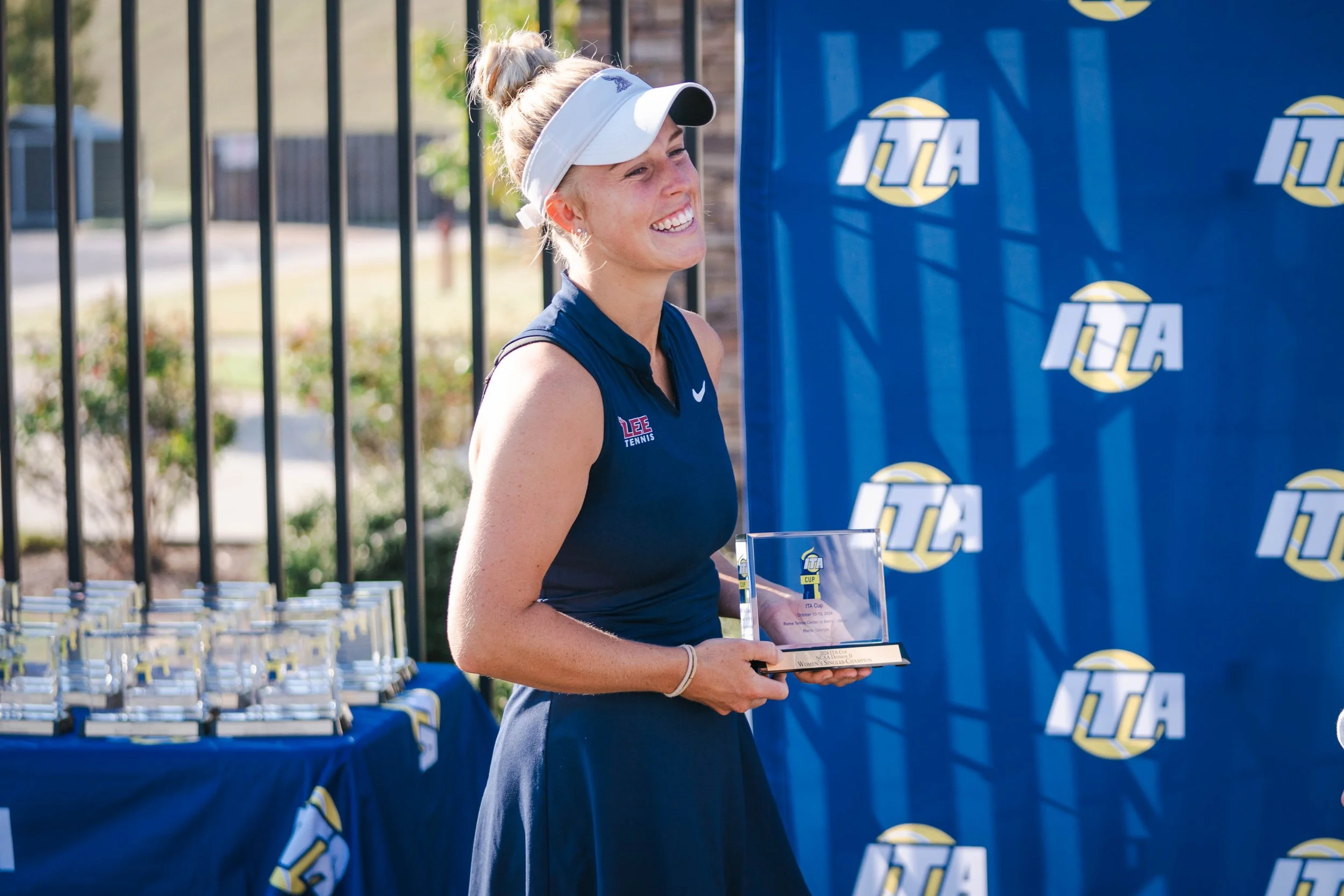 A young woman wearing a navy blue athletic uniform and a white visor, smiling while holding a trophy at an outdoor awards ceremony for an ITA tennis tournament, with a blue backdrop and a table of trophies behind her.