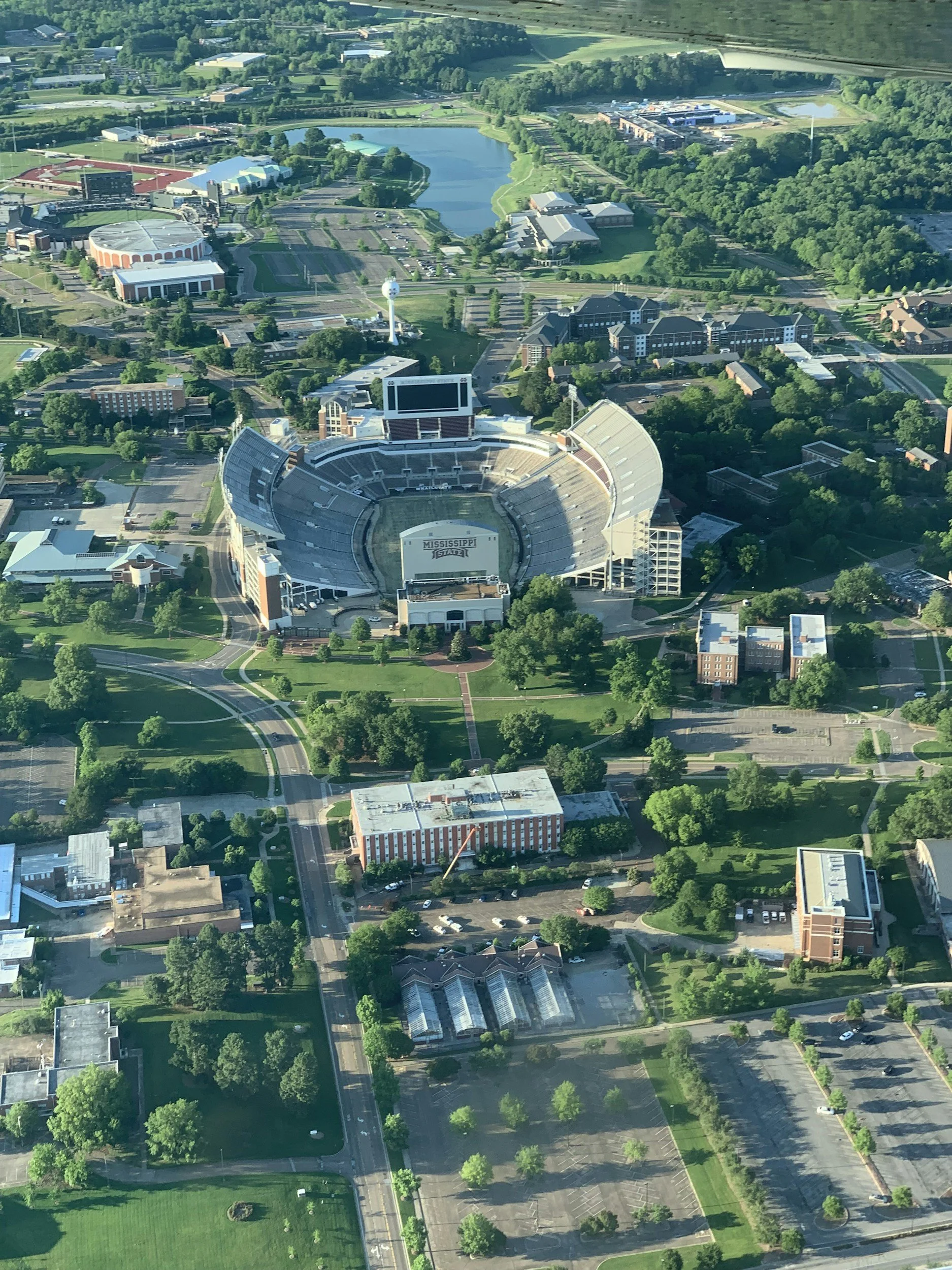 Aerial view of Mississippi State University campus, showing a large stadium, a lake, parking lots, roads, and surrounding greenery.