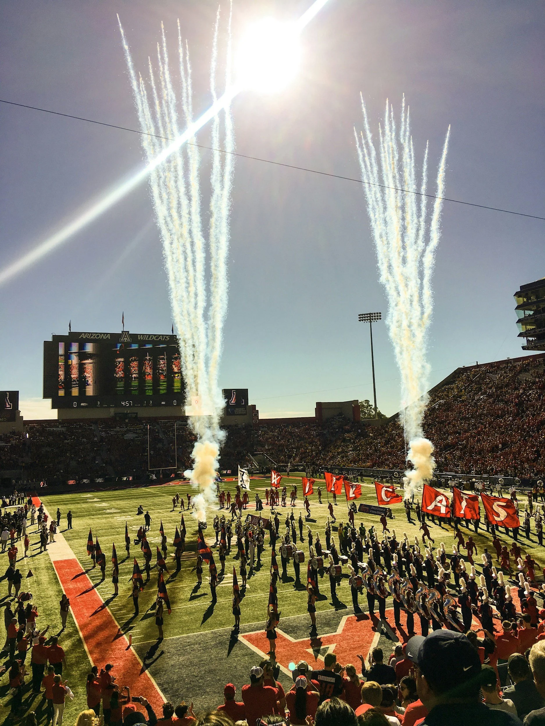 A football game halftime show with fireworks in the sky, a marching band on the field, and fans watching from the stands.
