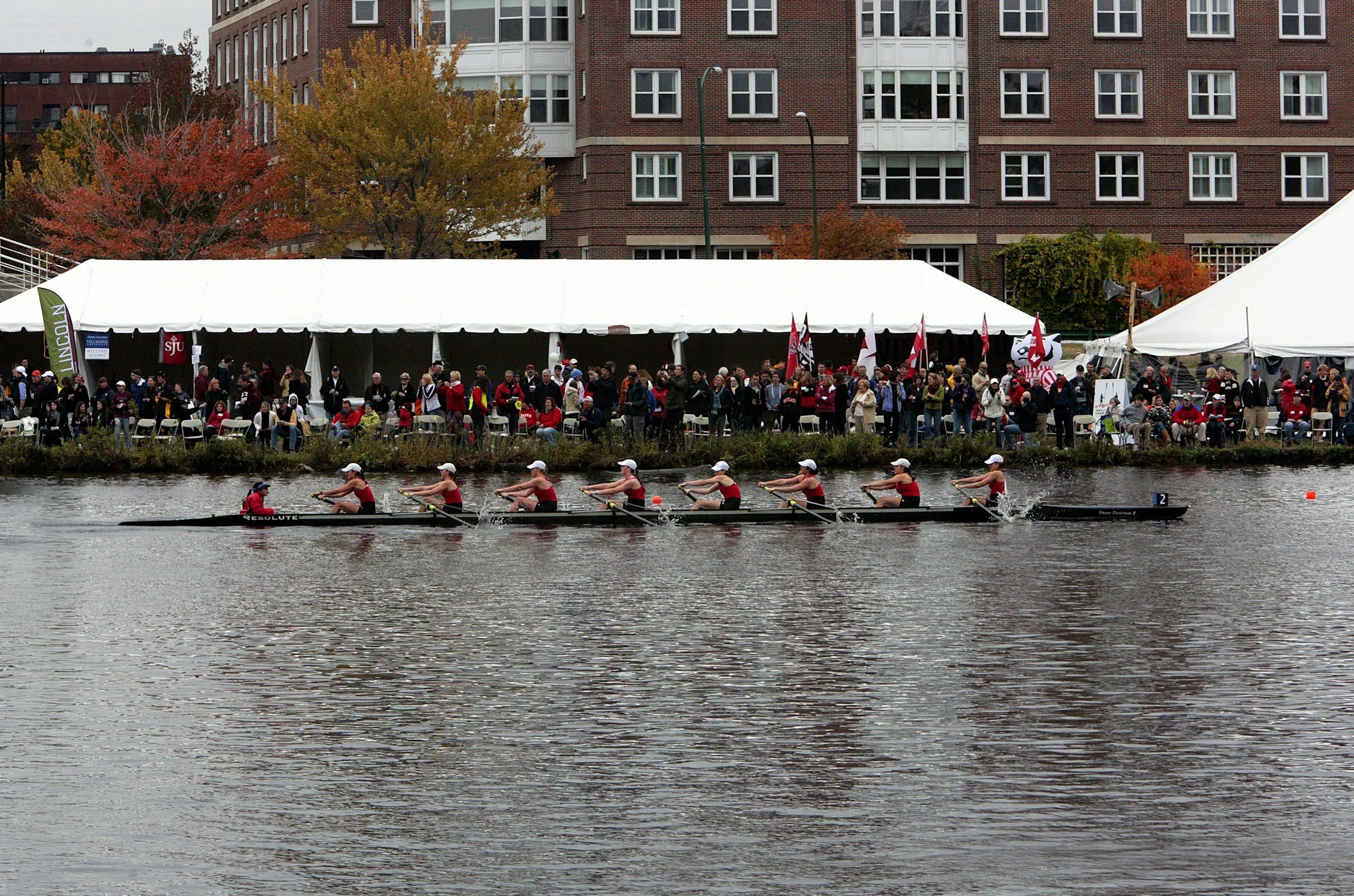 A rowing team of eight people in red and black uniforms on a river, with a row of tents and an apartment building with red brick and white window frames in the background, and autumn trees with orange and red leaves.
