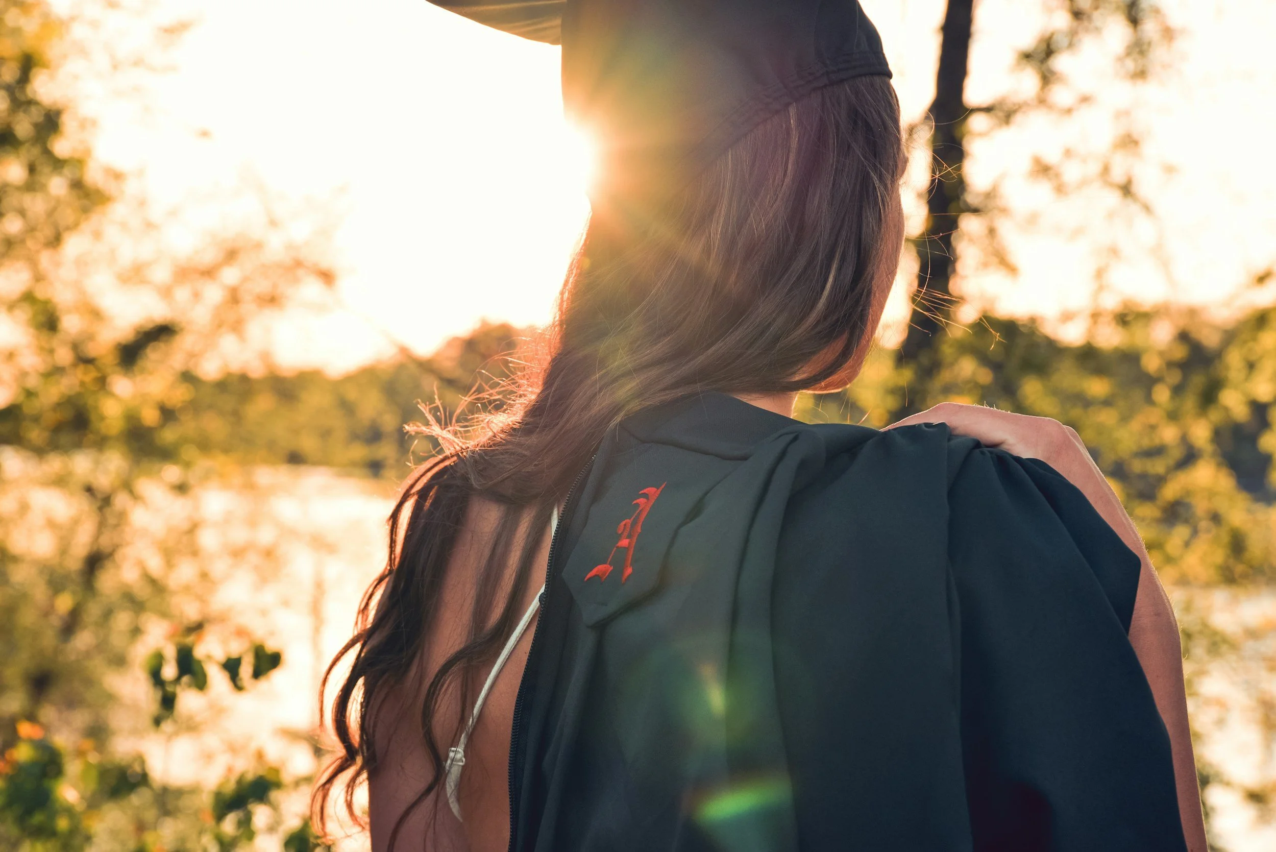 A woman with long brown hair wearing a black jacket and a black cap standing outdoors in the sunlight during sunset, facing away from the camera, with trees and blurred autumn leaves in the background.