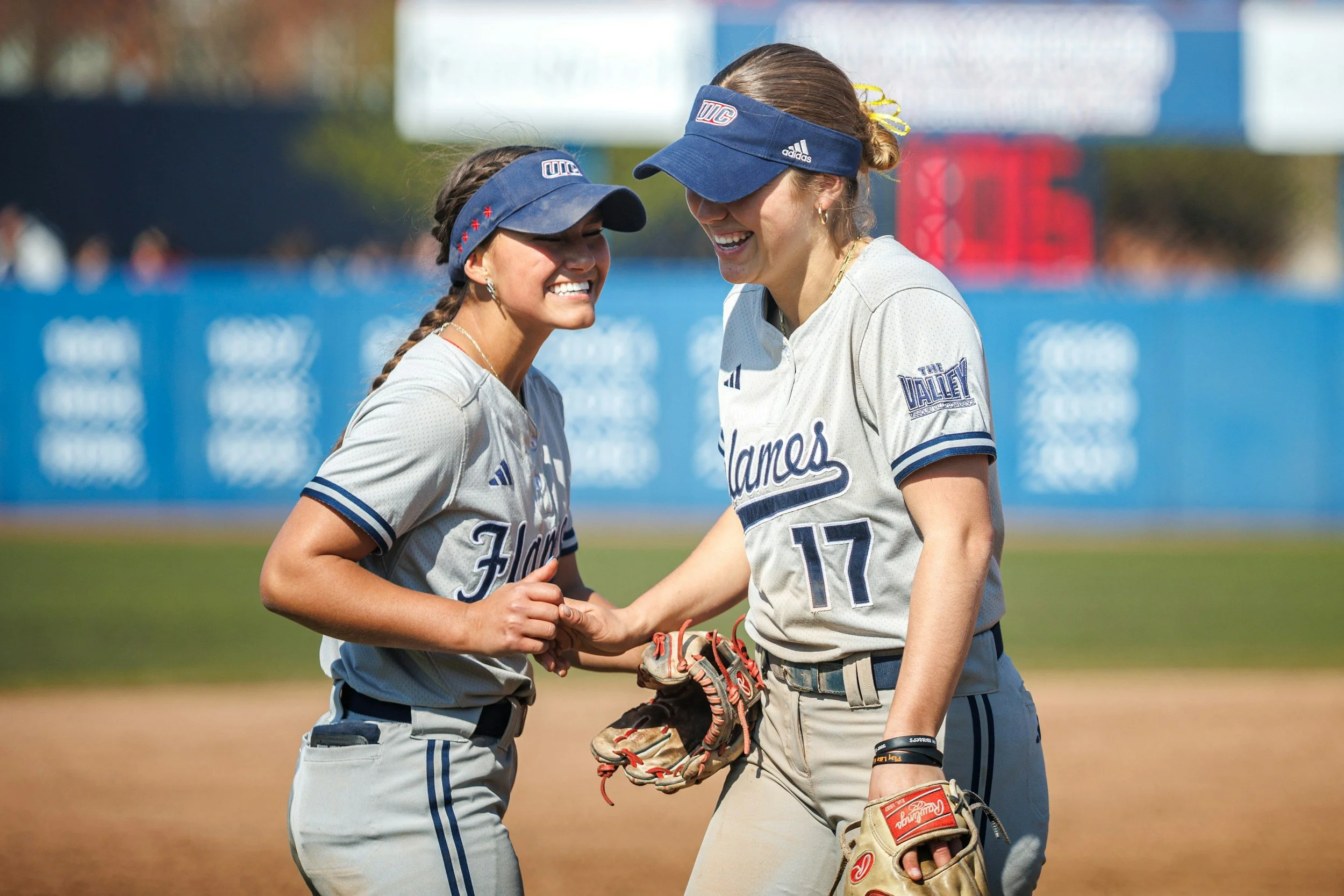 Two female softball players in uniform, smiling and shaking hands on the field.