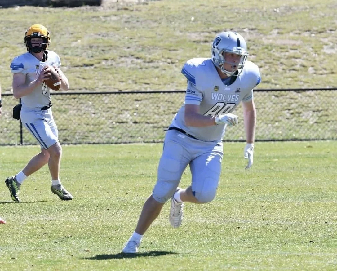 Two football players from the Wolves team practicing on the field, one running with the ball and the other preparing to catch or block, with a grassy area and a fence in the background.