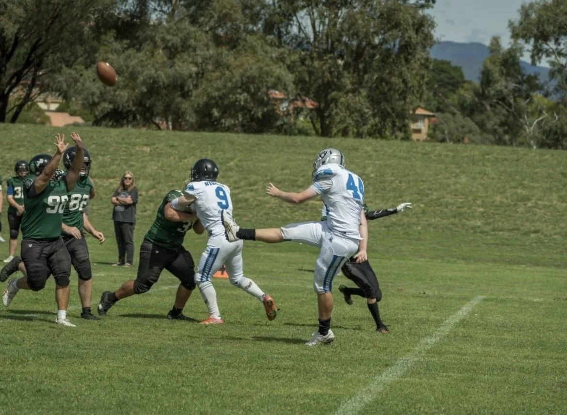 Football players in action during a game, with one player kicking the ball and others trying to block or catch it on a grassy field.