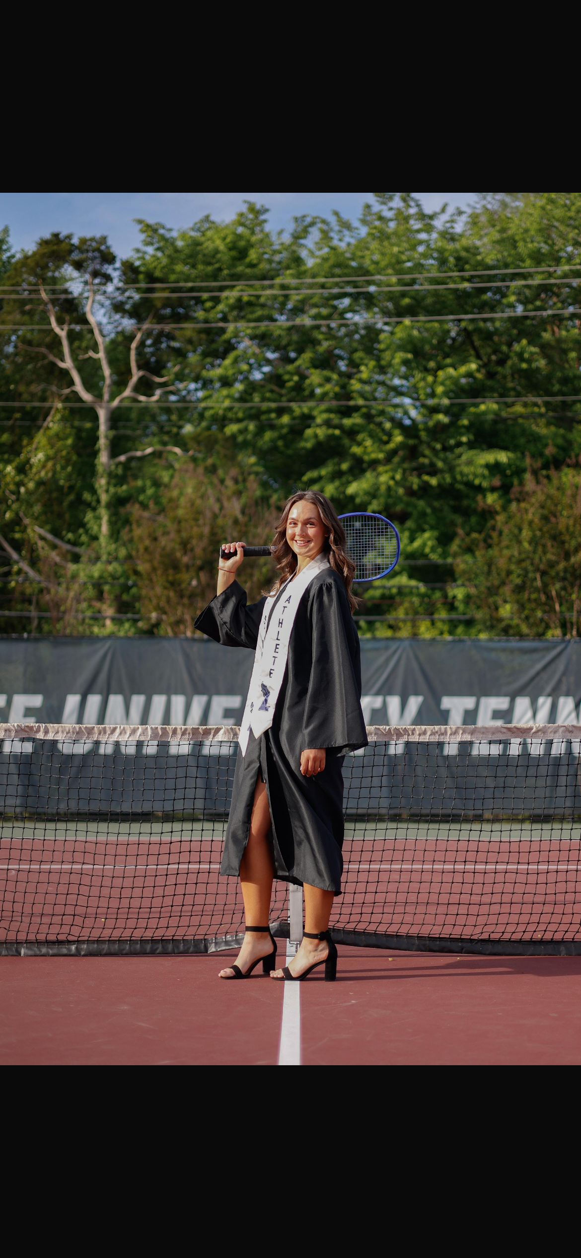 A young woman in a graduation gown and heels standing on a tennis court with a tennis racket over her shoulder, smiling at the camera.