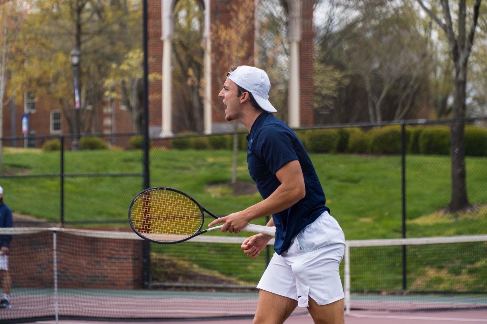 A young man wearing a navy blue shirt, white shorts, and a white backwards cap playing tennis on an outdoor court with a green grassy background and trees.