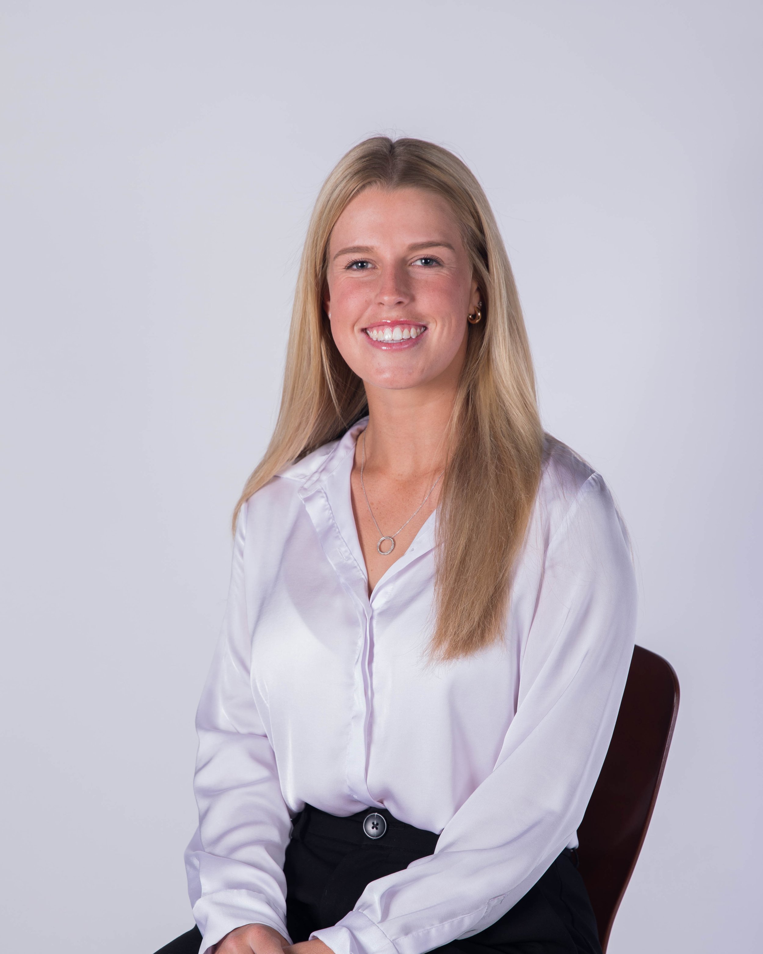 Portrait of a young woman with long blonde hair, wearing a white button-down shirt and black pants, sitting on a chair against a plain light gray background, smiling.