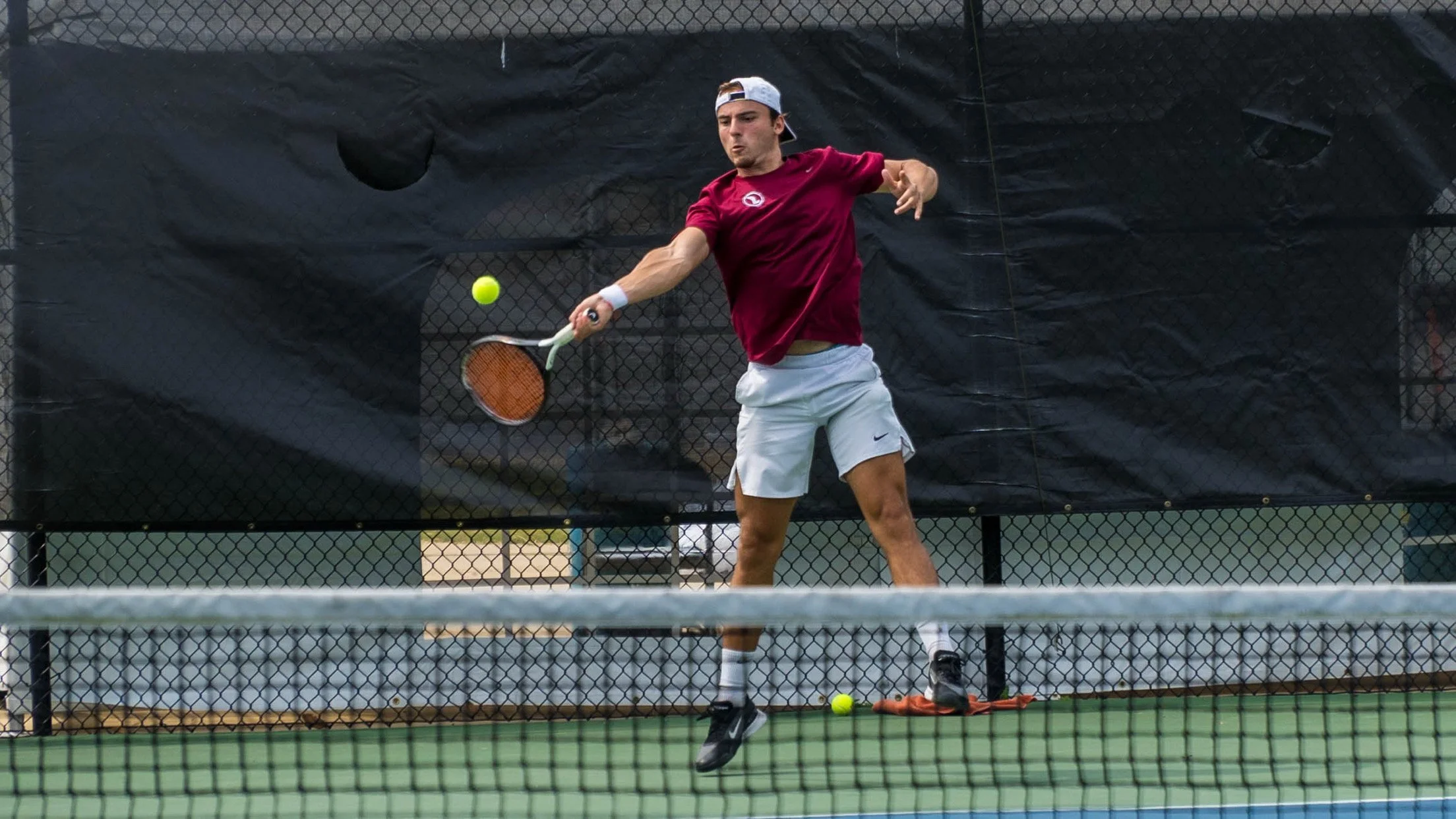 A young man playing tennis on a court, wearing a red shirt, white shorts, and a white cap, hitting a tennis ball with a racket.