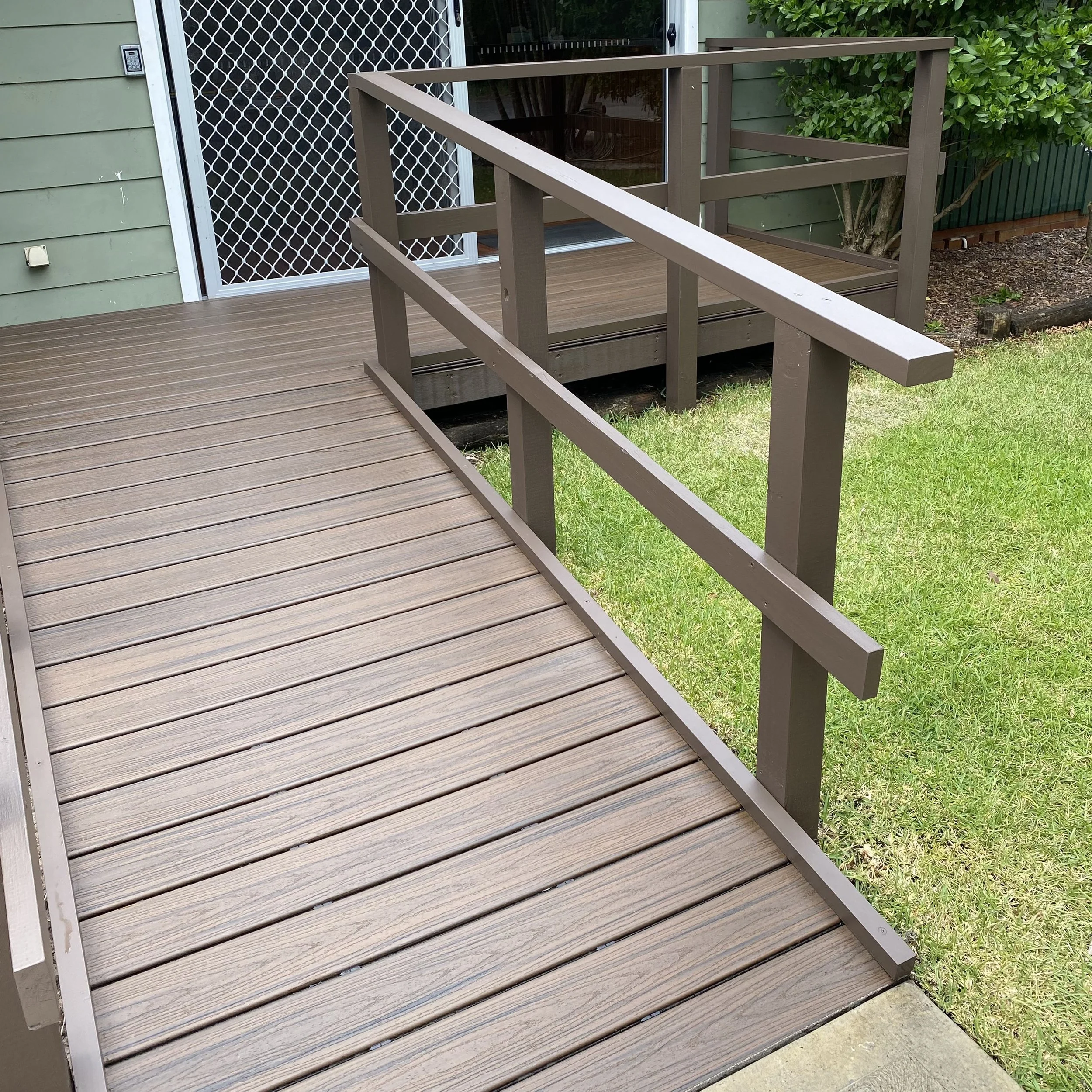 A composite deck entry deck complete with a railing outside a house, featuring a sliding glass door with a security screen. The house has green siding, and there is a garden with bushes and grass nearby.