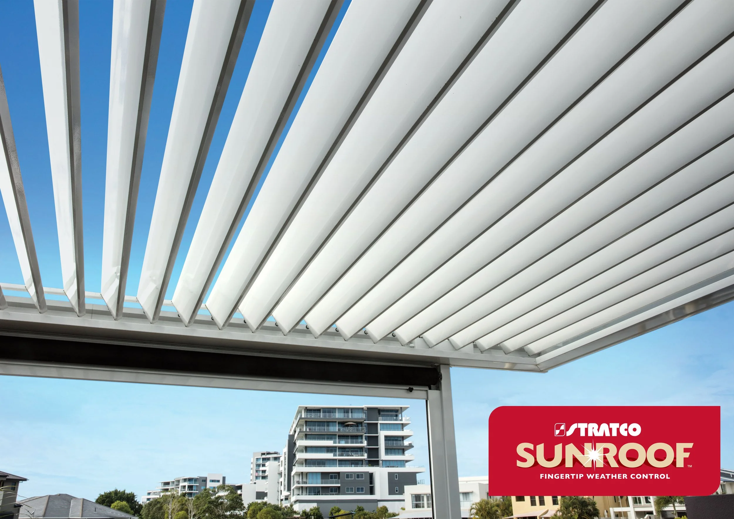 View from a balcony showing a white louvered pergola roof, modern high-rise buildings, and a blue sky with some clouds, with a red advertisement banner for Stratco Sunroof in the bottom right corner. Platinum Decks authorised installer.