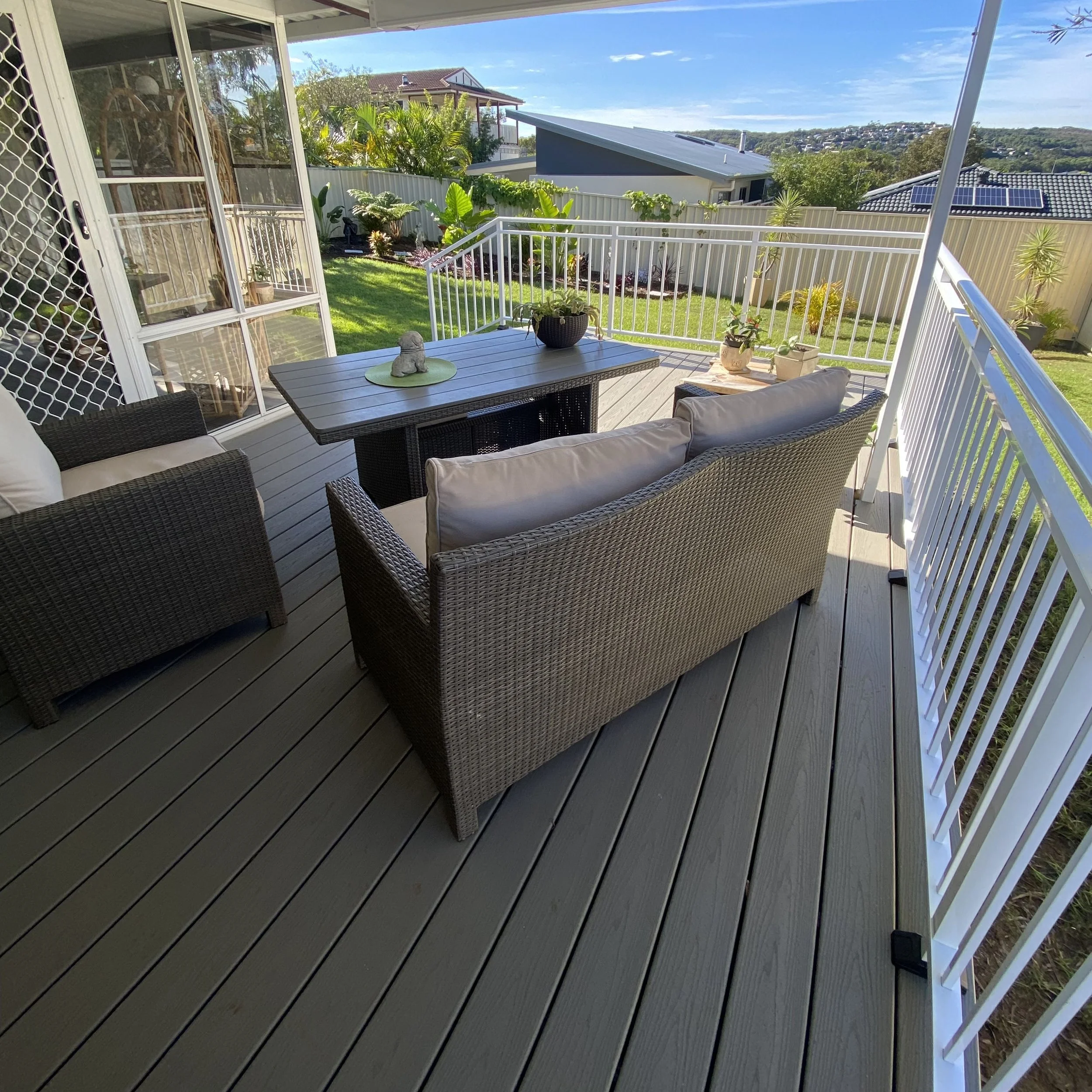 A backyard patio with a table, chairs, and potted plants, enclosed by a white balustrade, Trex deck under a blue sky.