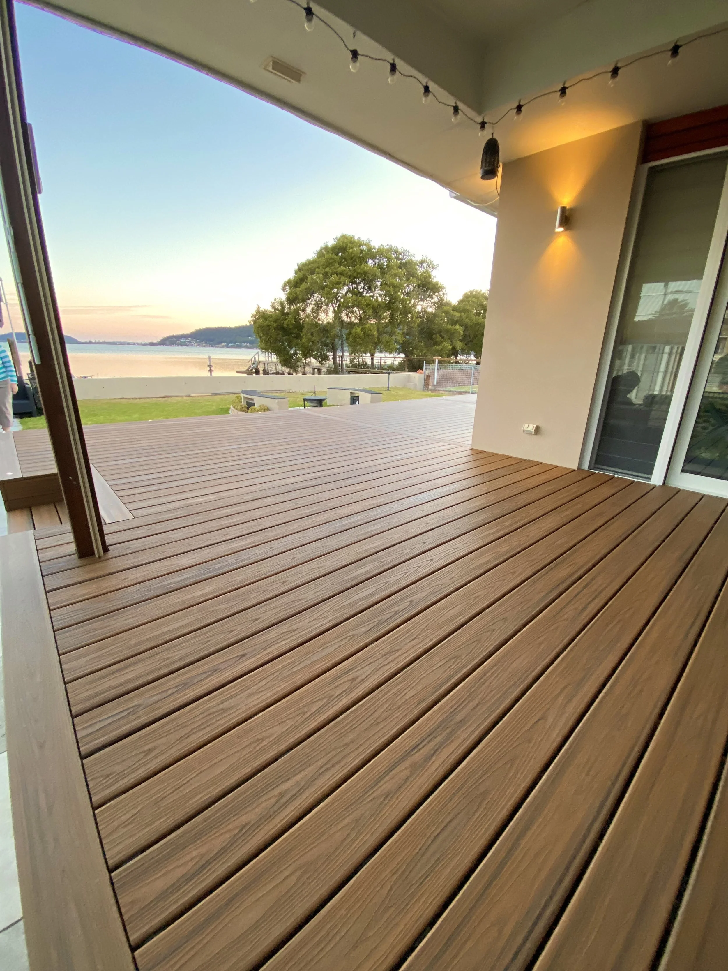 View of a balcony with stylish low maintenance composite decking, overlooking trees, a body of water, and a sunset, with outdoor lighting and string lights hanging from the ceiling.