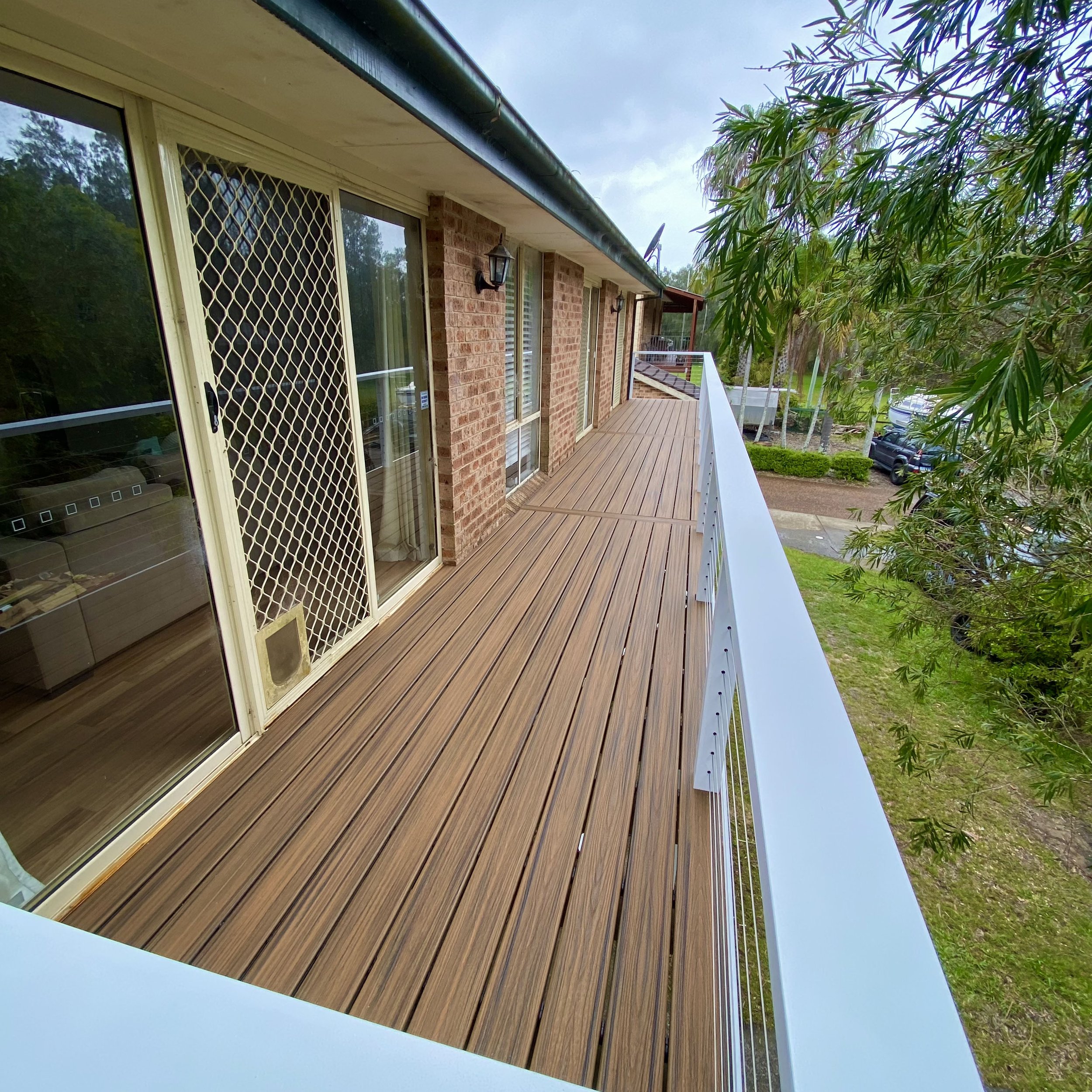 View of a house's exterior deckj with sliding glass doors, brick walls, outdoor lighting, and a new long composite deck, surrounded by trees and parked cars in the background.