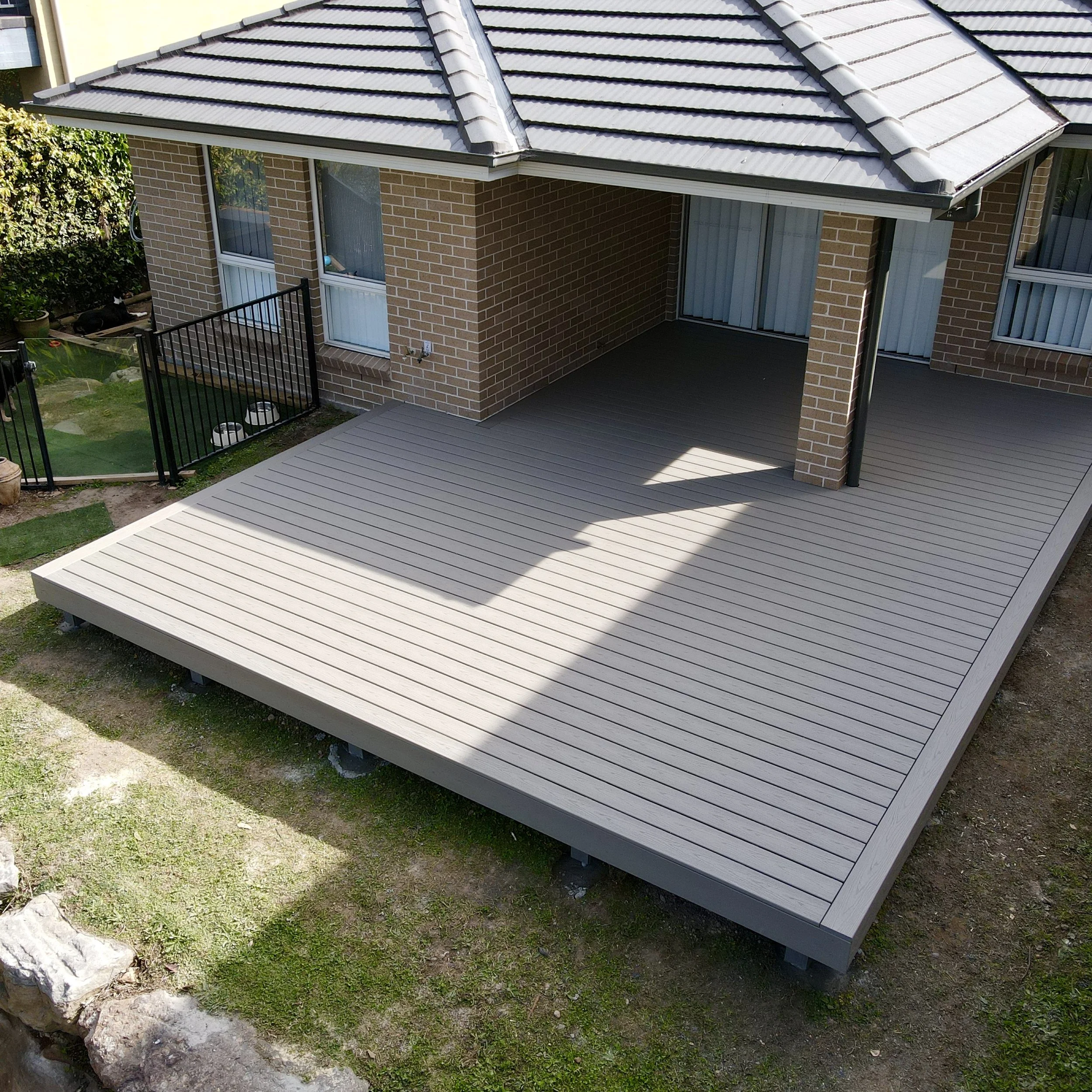 A two-story brick house with a covered outdoor deck area. The deck has white wooden flooring and a black metal railing. The house has large windows with drapes, and the roof is made of gray tiles. There is a small garden with greenery and rocks surro