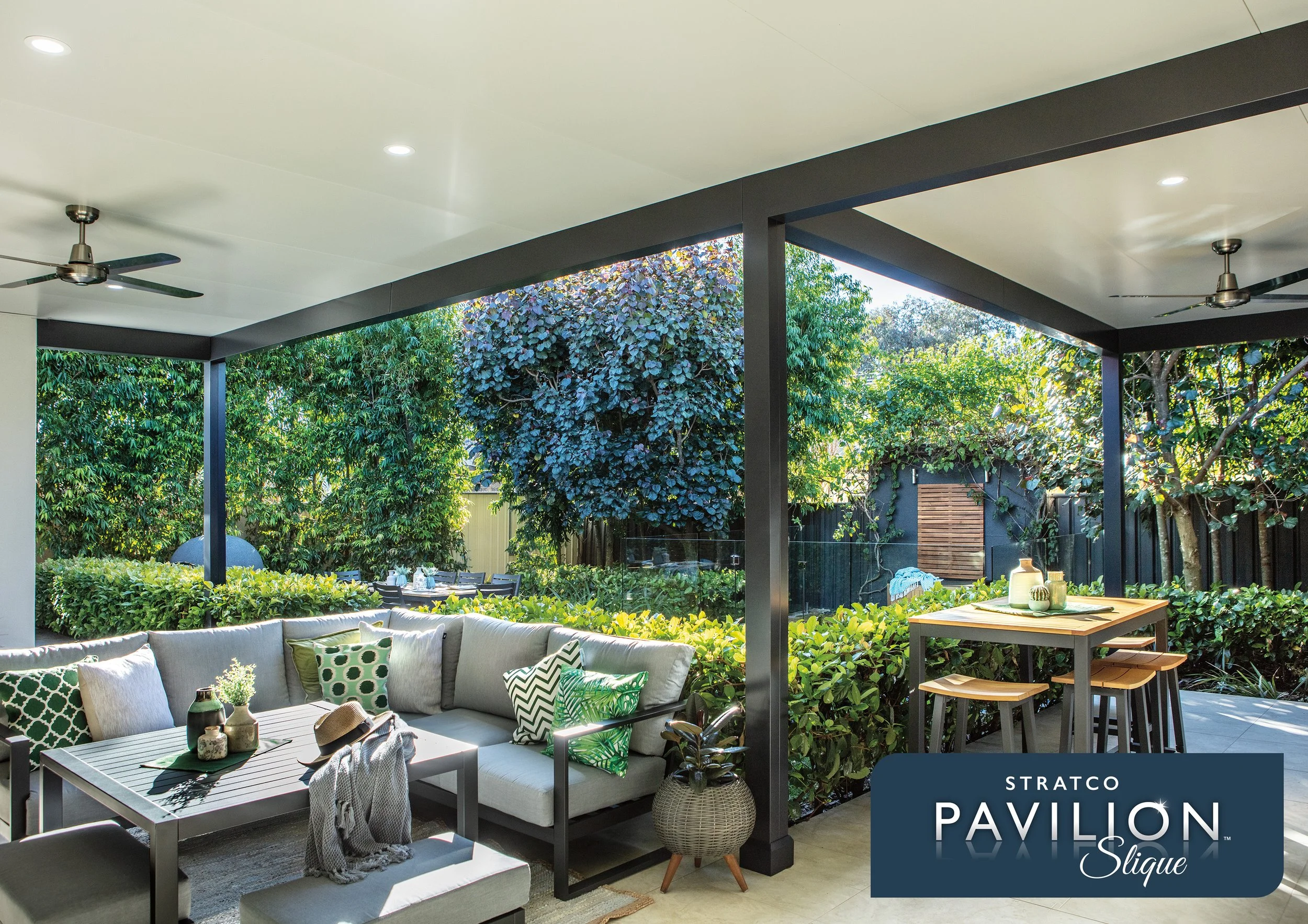 A modern outdoor patio with a beige sectional sofa, patterned cushions, a wooden coffee table with decorative items, a dining table with chairs, lush green trees and shrubs surrounding the space, a black metal frame pergola, ceiling fans, and built-i